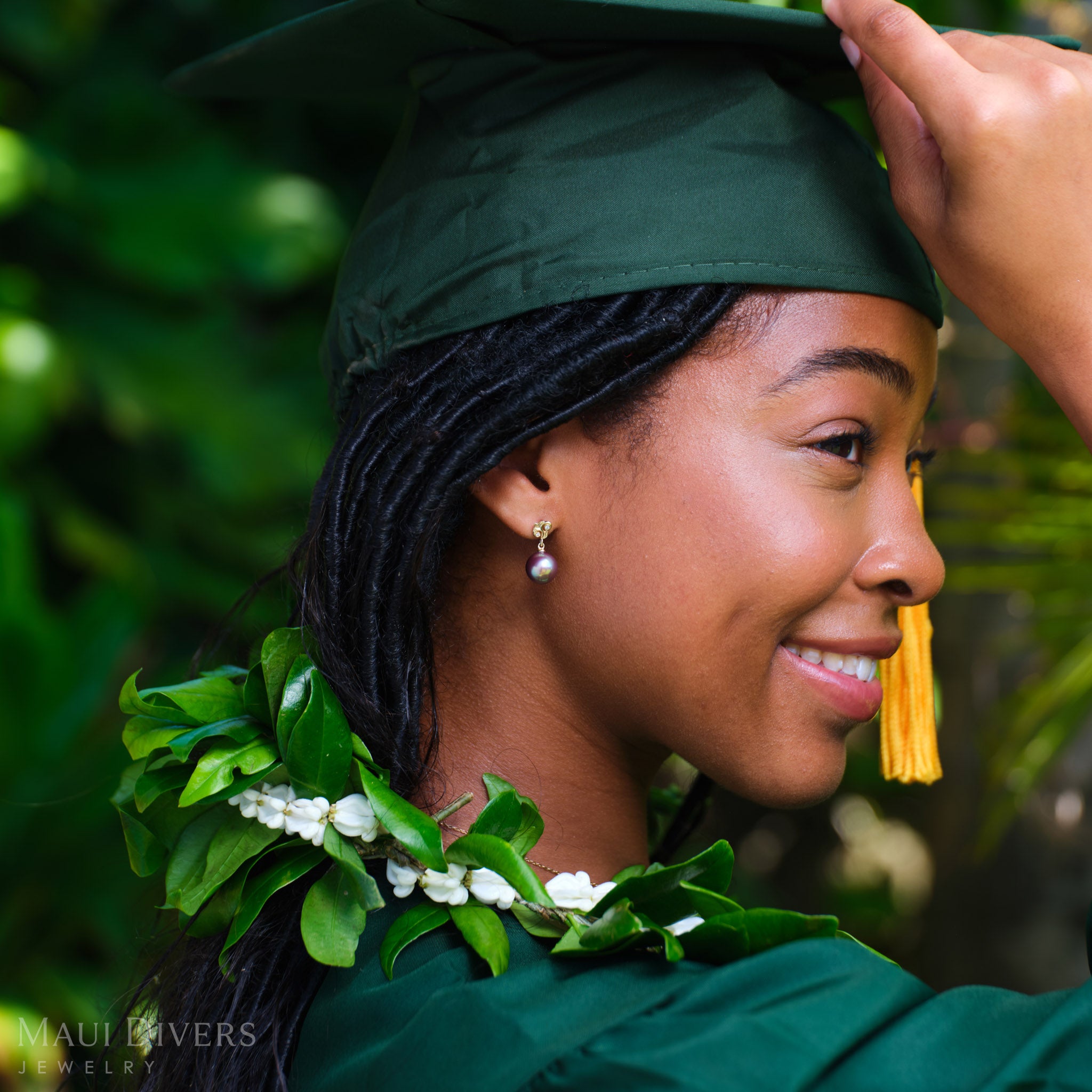 Smiling woman in a green graduation cap and gown wearing a Living Heirloom Freshwater Pearl Earring in 14k yellow gold with diamond accents on her right ear, against a blurred green background