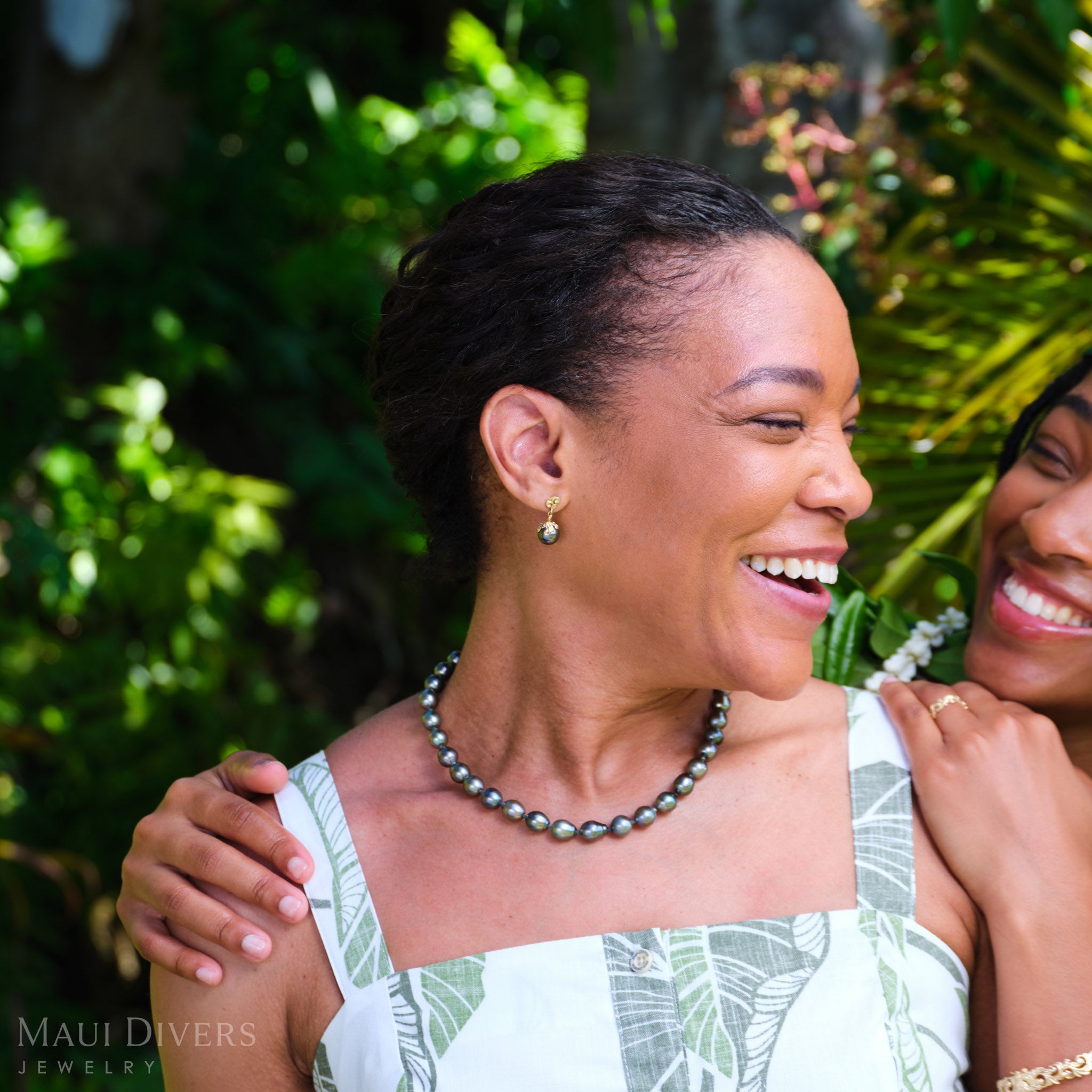 Smiling woman wearing a Living Heirloom Tahitian black pearl earring in 14k yellow gold with diamond accents on her right ear, and a Tahitian black pearl strand necklace, looking toward another smiling woman against a blurred forest background