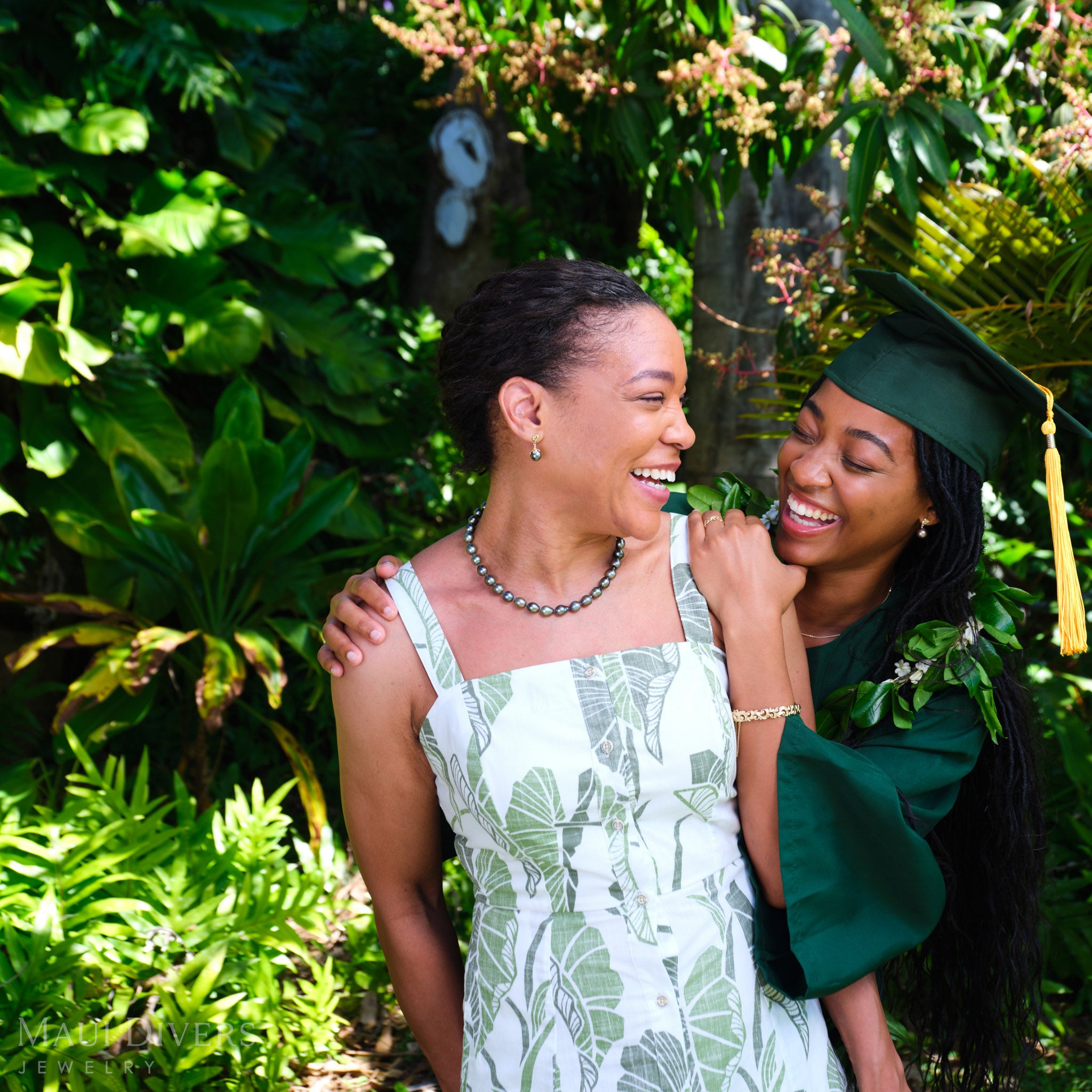Two women smiling at each other, one wearing a Living Heirloom Tahitian black pearl earring in 14k yellow gold with diamond accents on her right ear and a Tahitian black pearl strand necklace, the other in a green graduation cap and gown with a Living Heirloom Hinge Bracelet on her left wrist
