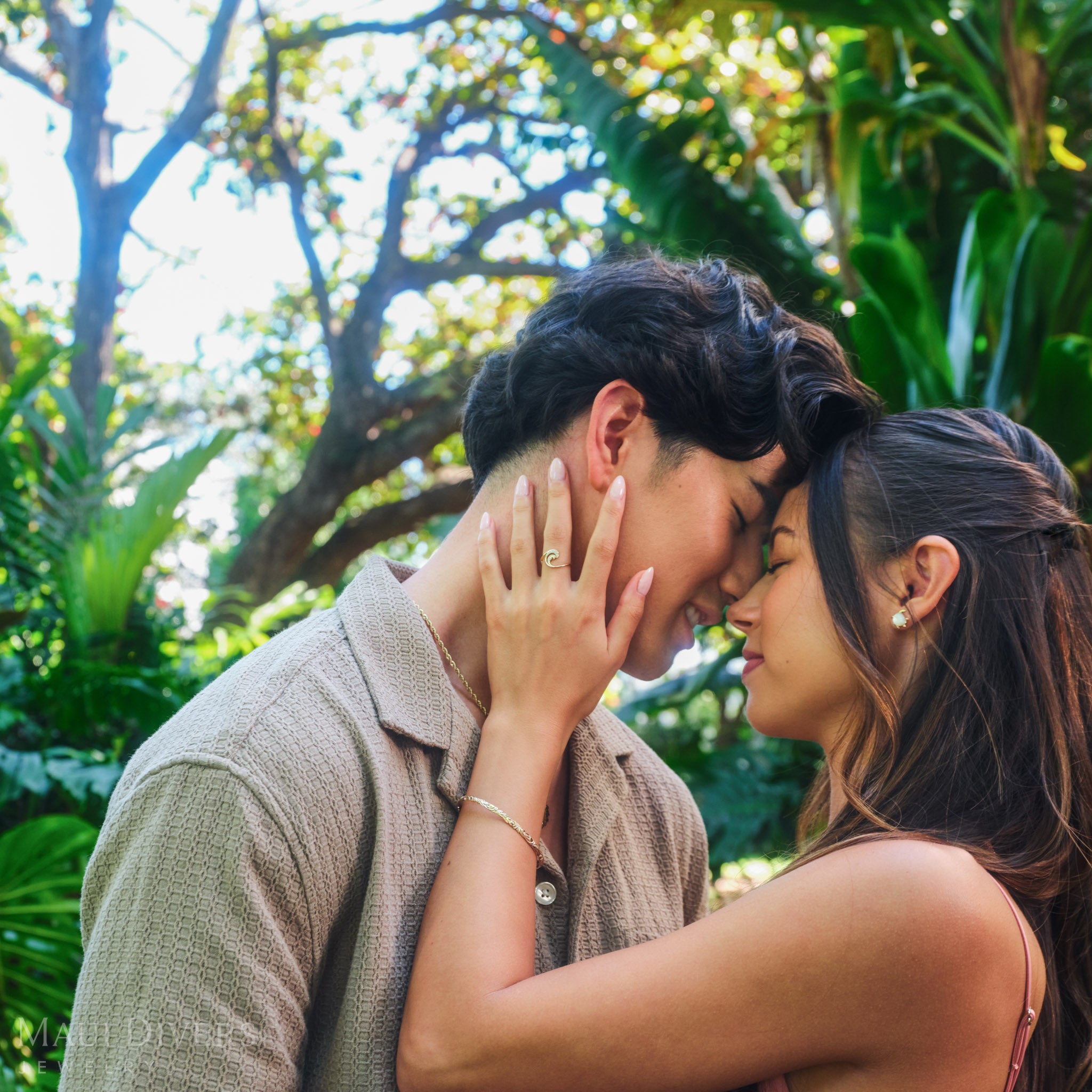 Smiling couple embracing, with the woman wearing a Hawaiian Heirloom Old English Scroll Bracelet in 14k yellow gold and a Nalu Ring in 14k yellow gold with diamonds, against a blurred forest background