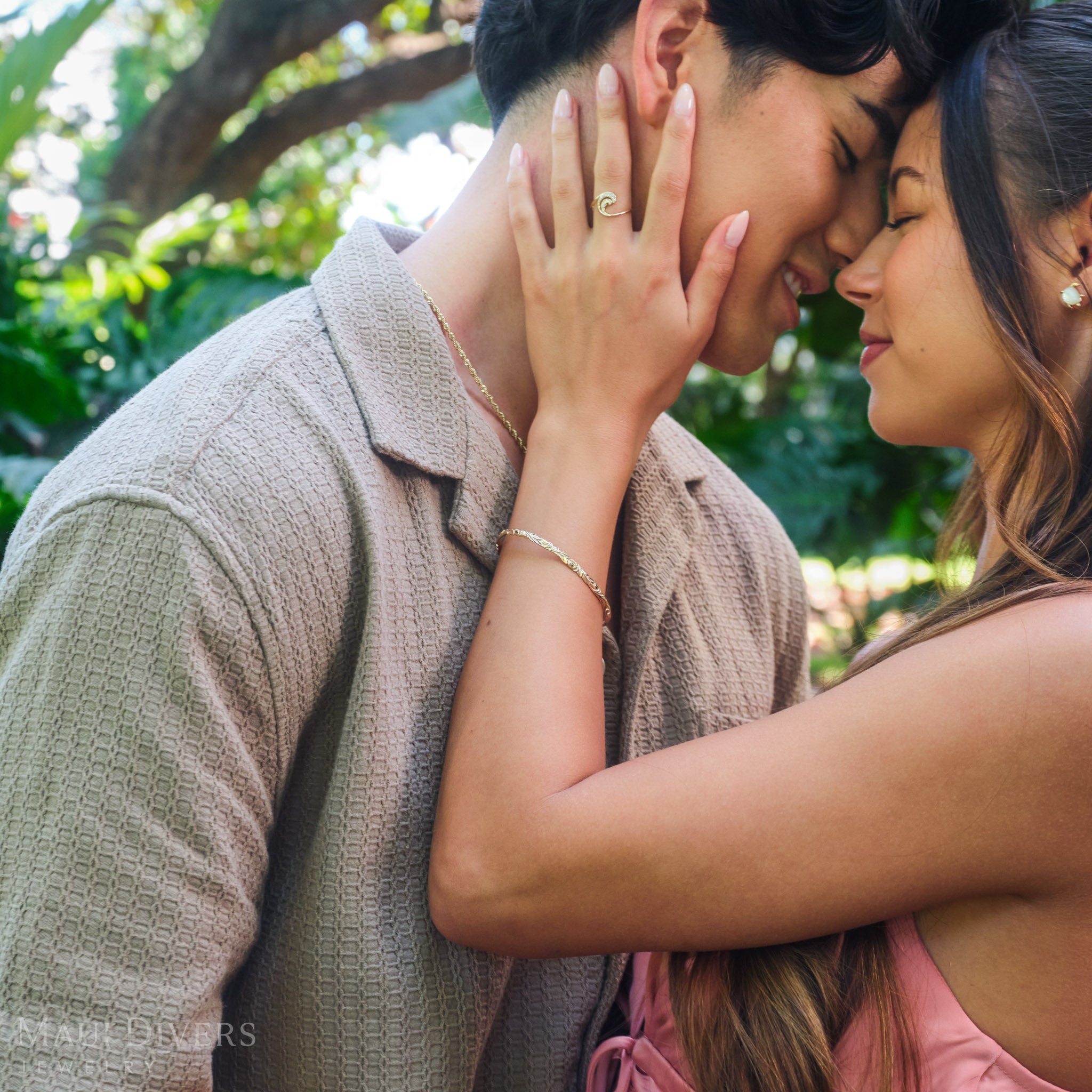 Close-up of a smiling couple embracing, with the woman wearing a Hawaiian Heirloom Old English Scroll Bracelet in 14k yellow gold and a Nalu Ring in 14k yellow gold with diamonds, against a blurred forest background