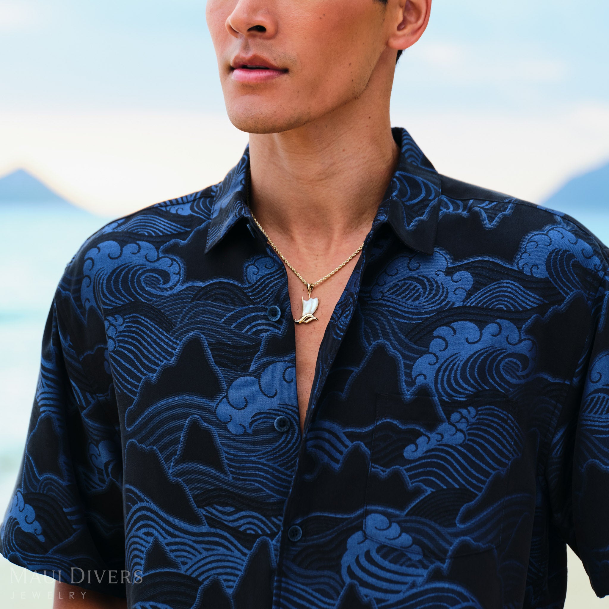 Waʻa (Canoe) Pendant on a gold chain worn by a man in a blue patterned shirt with mountains in the background.