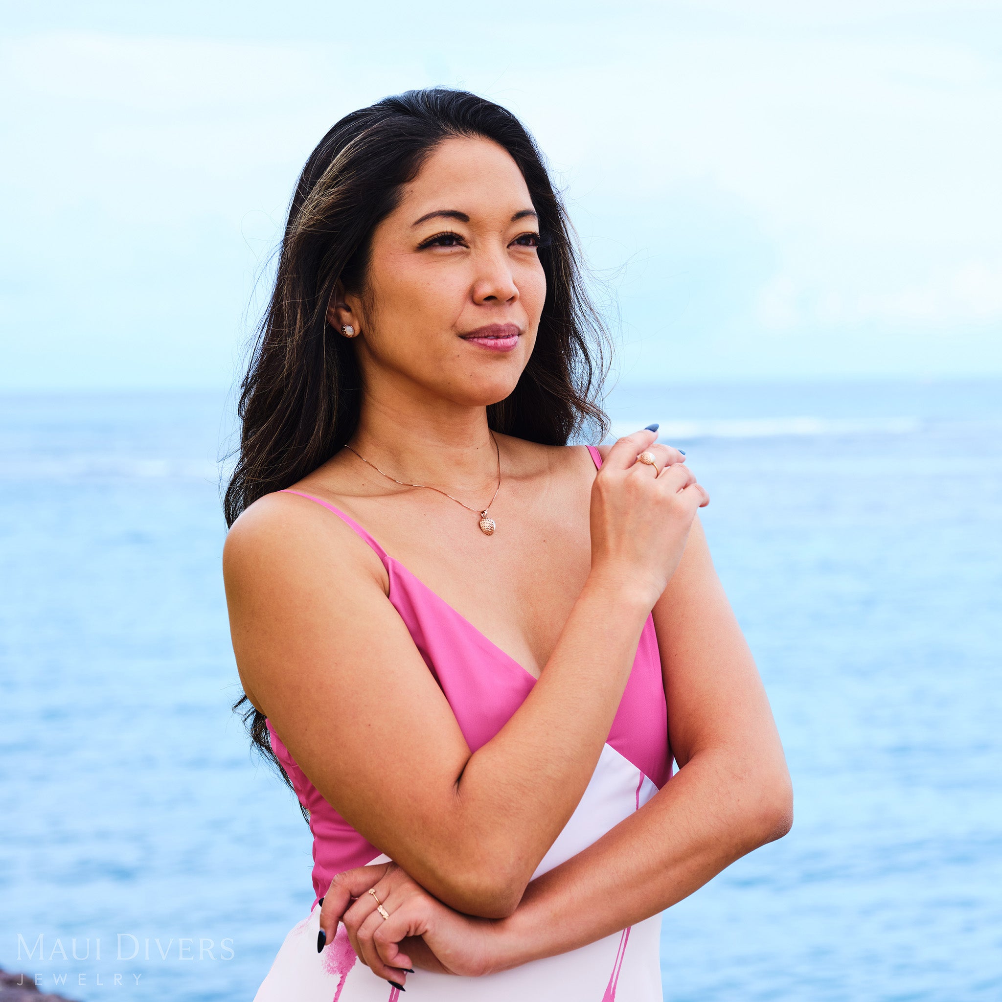 Smiling woman in a pink dress wearing a Cute Fruits Lychee Mother of Pearl Pendant in 14k rose gold around her neck with matching Cute Fruits Lychee Mother of Pearl Ring and Earrings, against a blurred ocean background