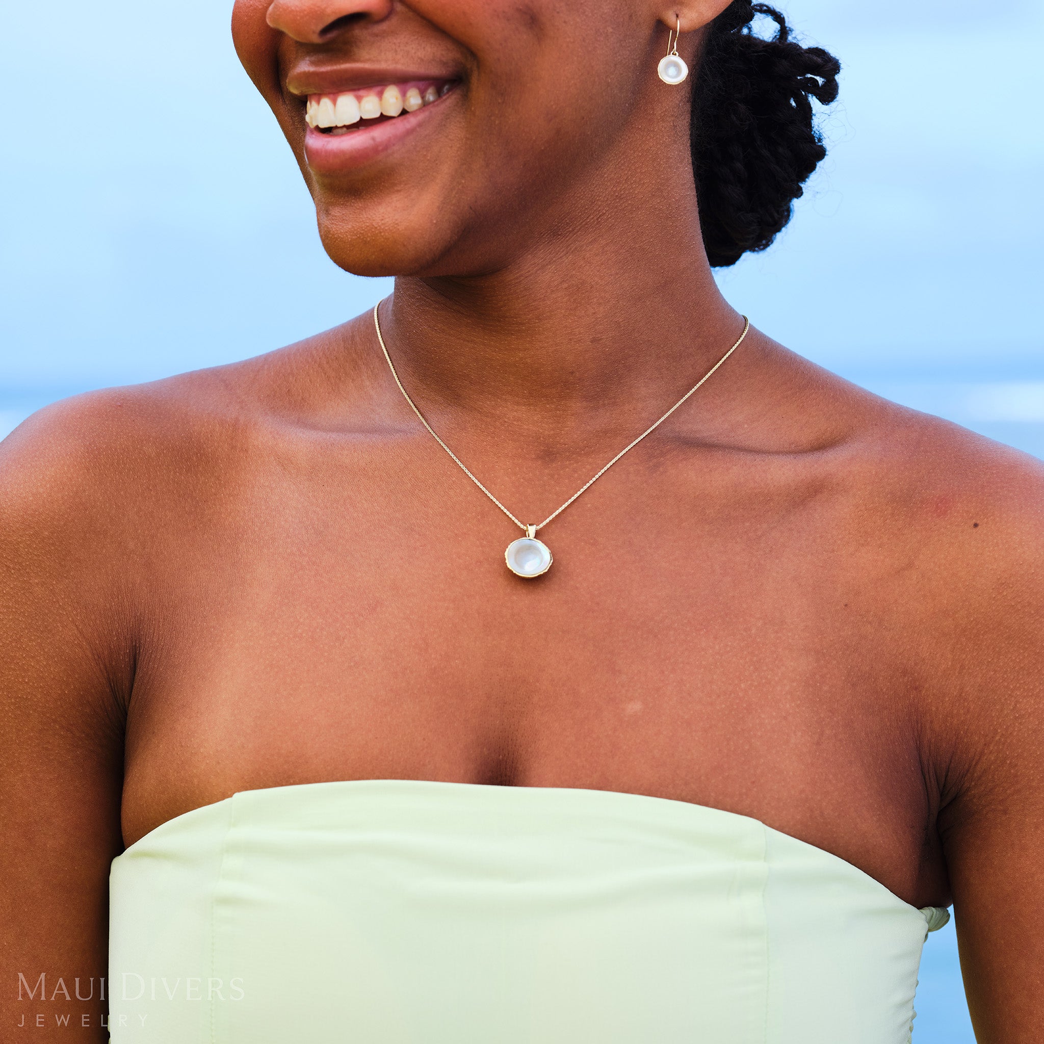 Close-up of a smiling woman in a pastel green dress wearing a Cute Fruits Coconut Mother of Pearl Pendant and a matching Cute Fruits Coconut Mother of Pearl Earring in 14k yellow gold