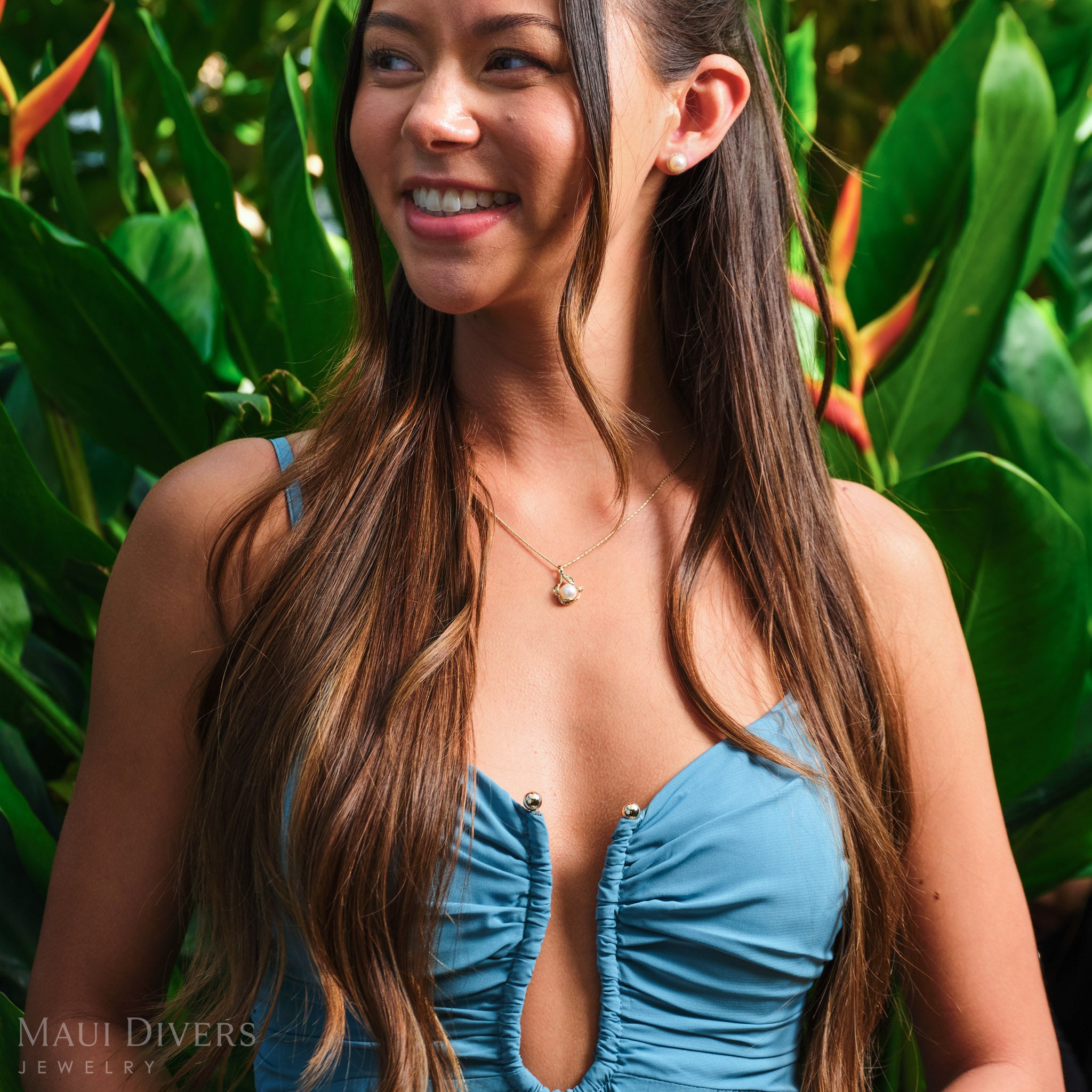 Smiling woman in a blue top wearing a Pick A Pearl Maile Pendant in 14k yellow gold with diamonds and a white pearl around her neck, against a blurred tropical background