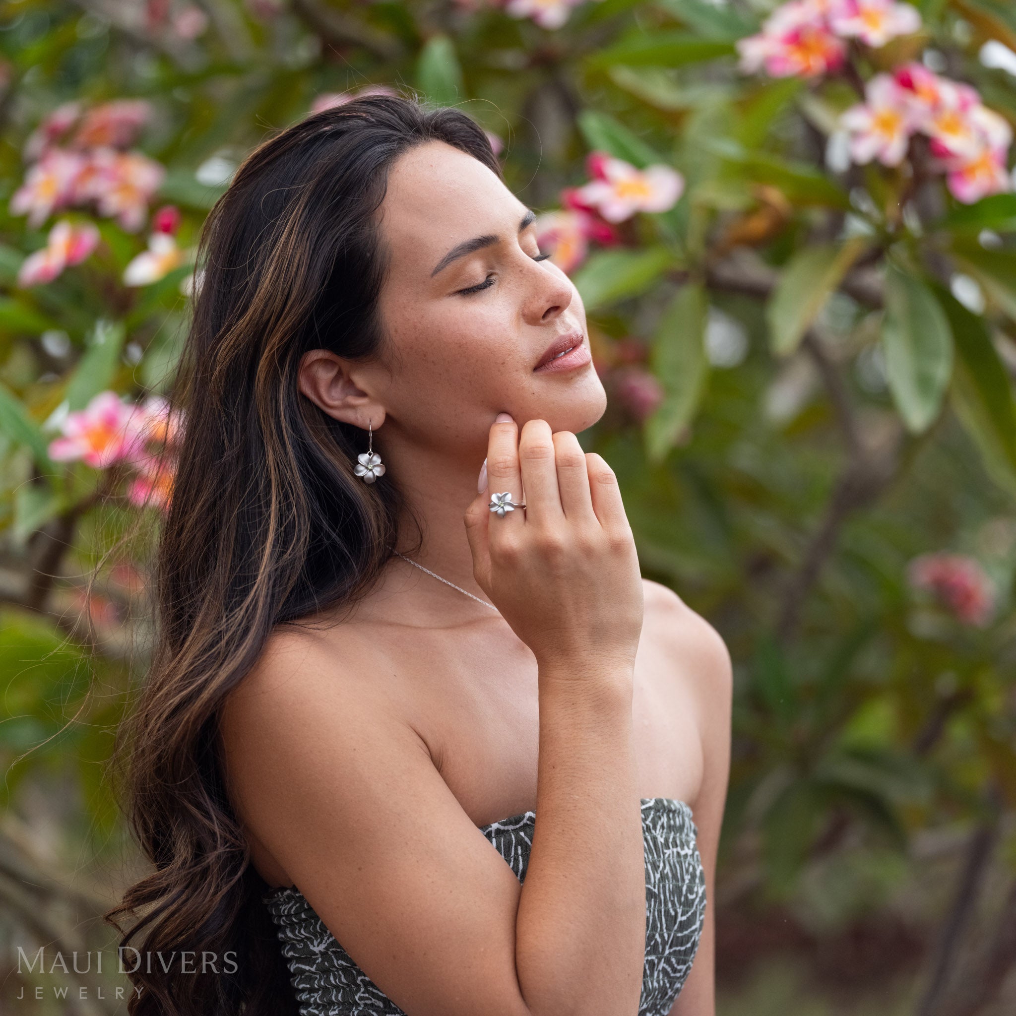 Woman closing her eyes wearing Plumeria Peridot Ring in Sterling Silver with matching earrings in front of a blurred plumeria tree with pink flowers and green leaves. 