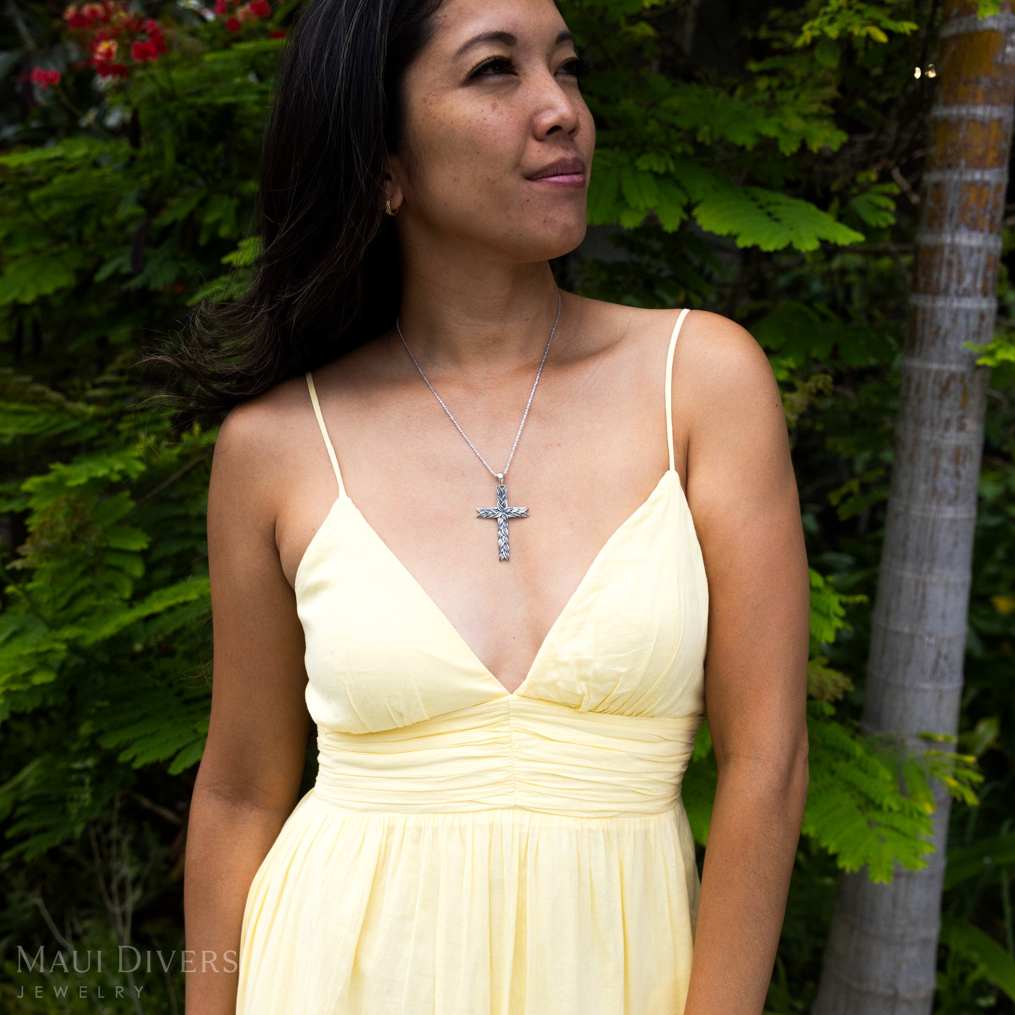 Woman in a yellow top wearing a Maile Cross Pendant in Sterling Silver against a leafy green background