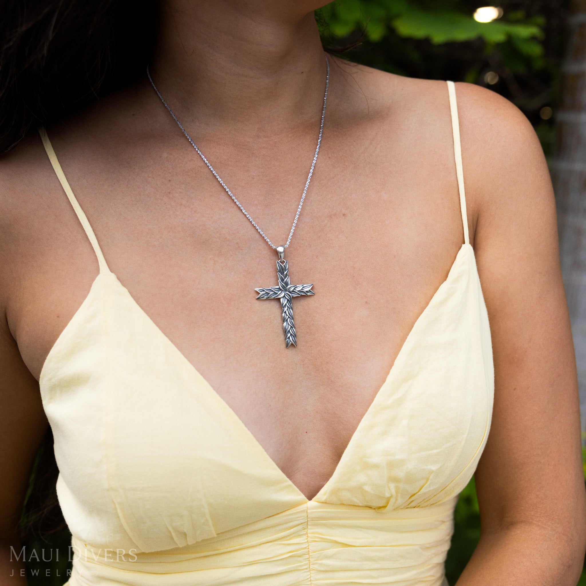 Close-up sideview of a woman in a yellow top wearing a Maile Cross Pendant in Sterling Silver against a blurred leafy green background