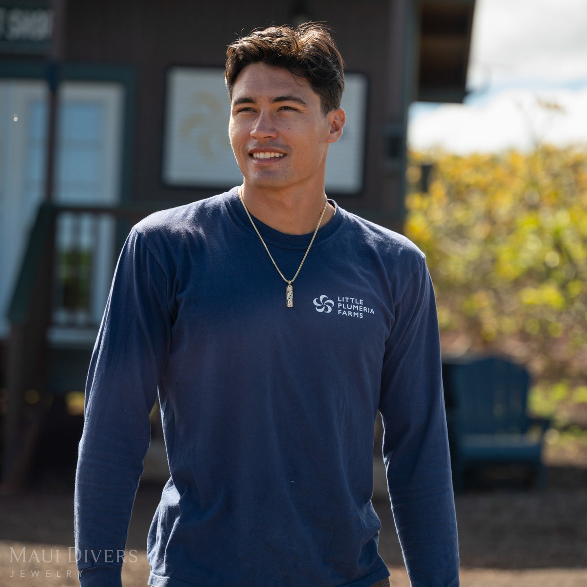 Dane Little wearing a gold vertical bar Hawaiian Heirloom pendant on a chain and a dark blue shirt with ʻLittle Plumeria Farmsʻ in white against a blurry house background with plumeria trees