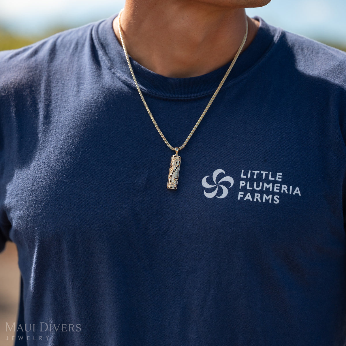 Close up shot of Dane Little wearing a gold vertical bar Hawaiian Heirloom pendant on a chain and a dark blue shirt with ʻLittle Plumeria Farmsʻ in white 