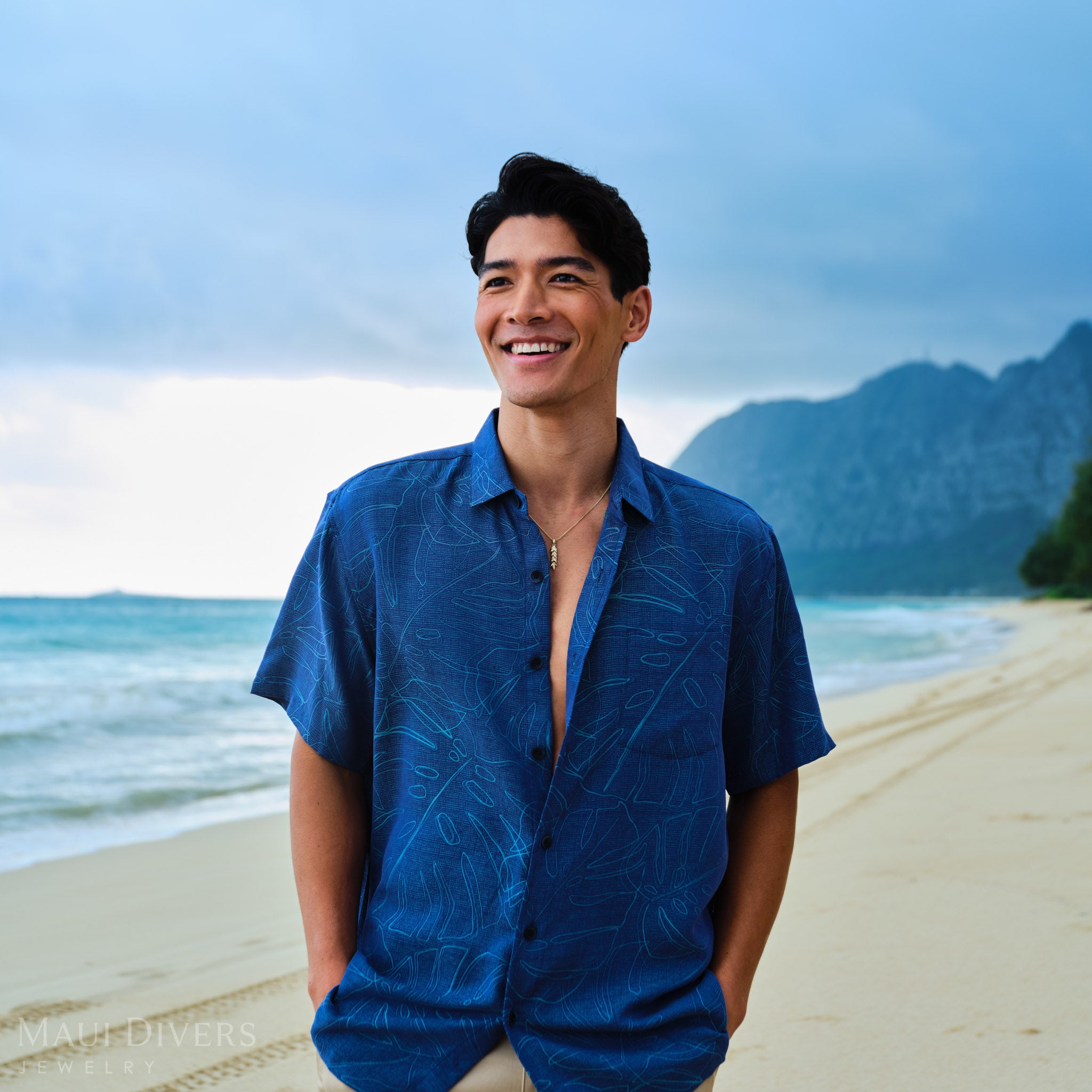 Man wearing a blue patterned shirt  with a gold maile leaf pendant on a chain on a beach with mountains in the background