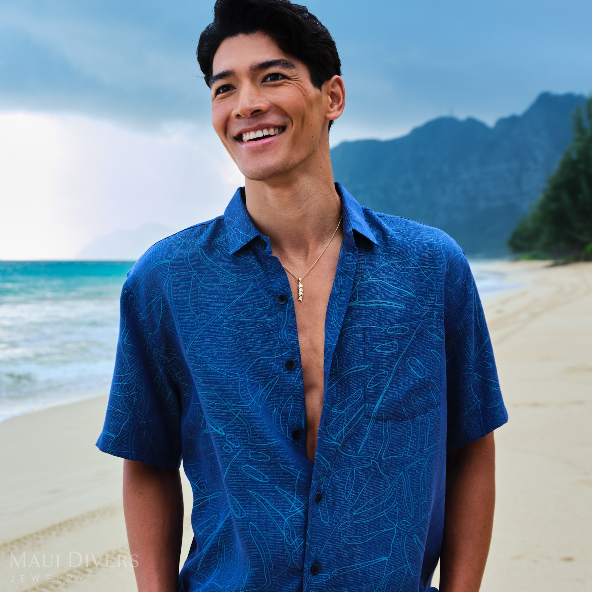 Man wearing a blue patterned shirt  with a gold maile leaf pendant on a chain on a beach with mountains in the background