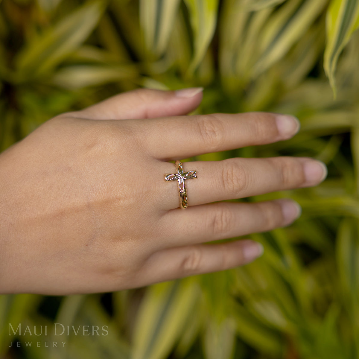 Close-up of a hand wearing a Hawaiian Heirloom Old English Scroll Cross Ring in 14k yellow gold, against a blurred leafy green background