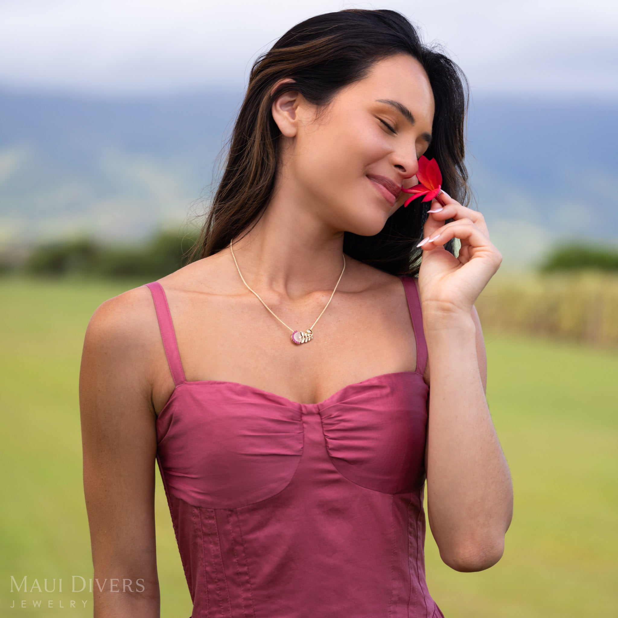 Woman smelling red flower wearing ʻŌhiʻa Lehua Ruby Pendant in Two Tone 14k Gold with Diamonds. 