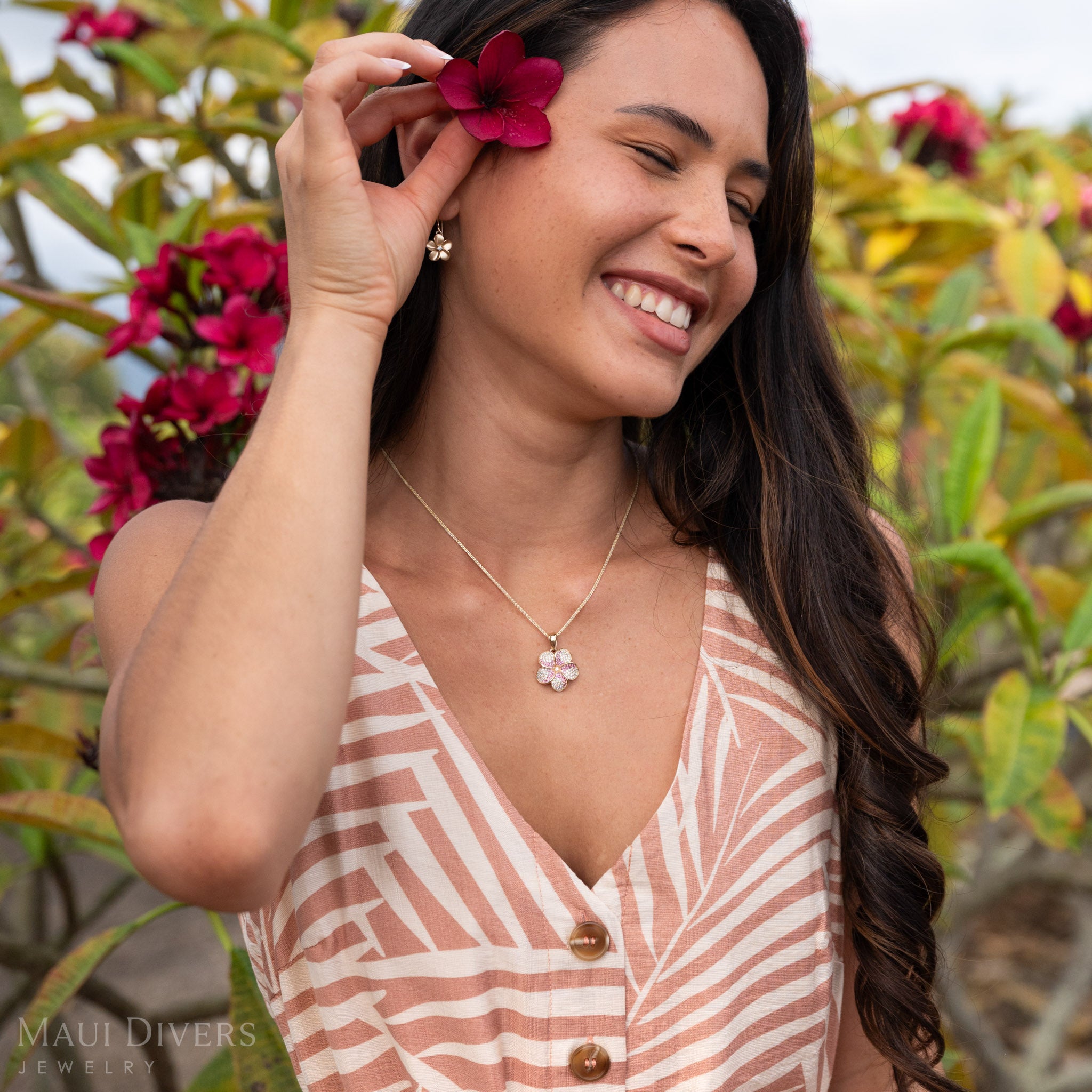 Woman putting a flower behind her ear wearing Plumeria Pink Sapphire Pendant in 14k Gold with Natural Diamonds and wearing the matching earrings.