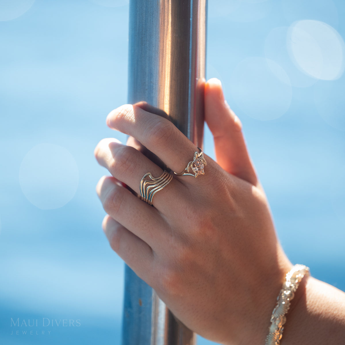 Close-up of a hand wearing a gold abstract wave ring and a sea turtle ring with plumerias against a blue sky background