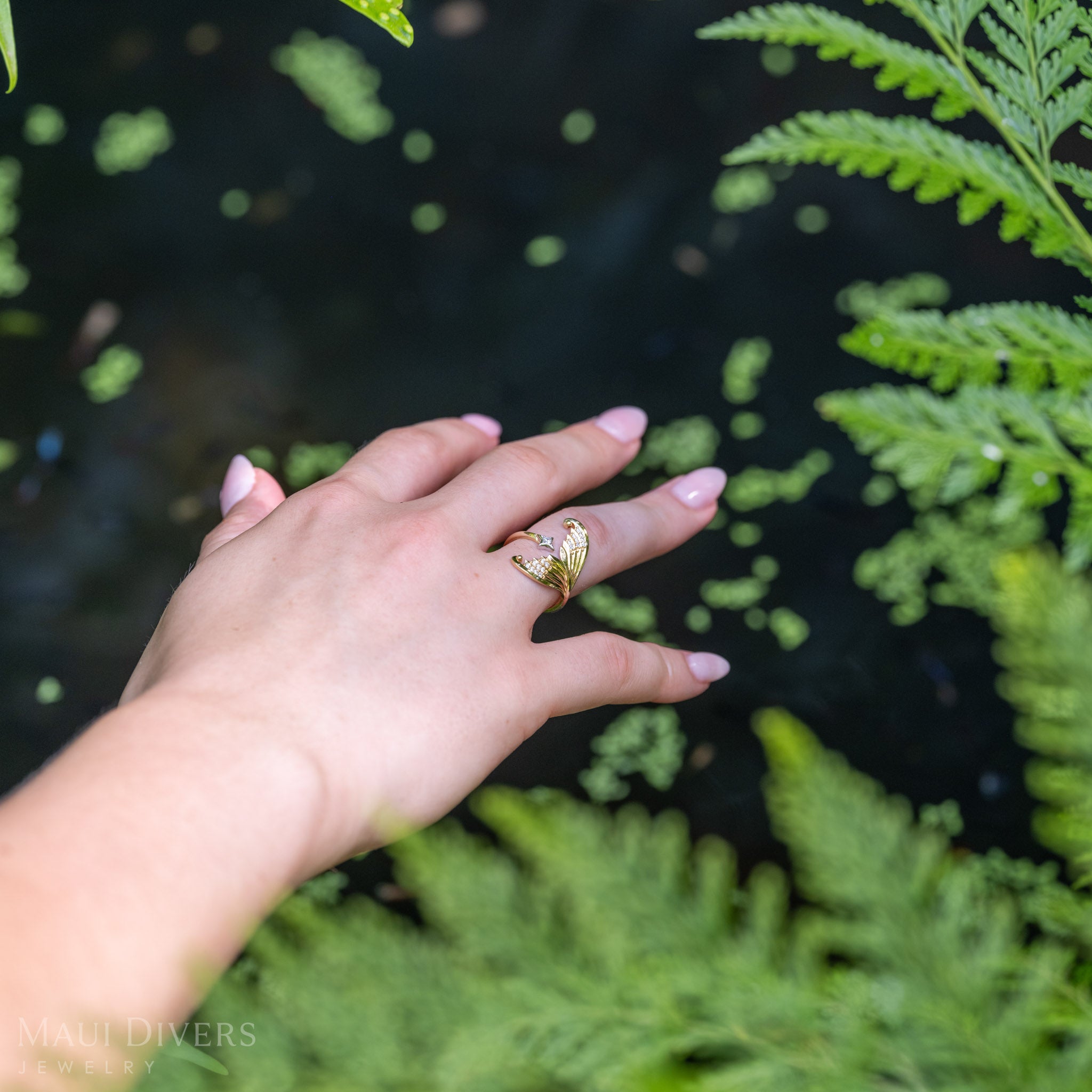 A 14k yellow gold diamond mermaid tail ring with accenting 14k white gold diamond star worn on a person's finger. The hand is positioned against a natural background of green ferns.