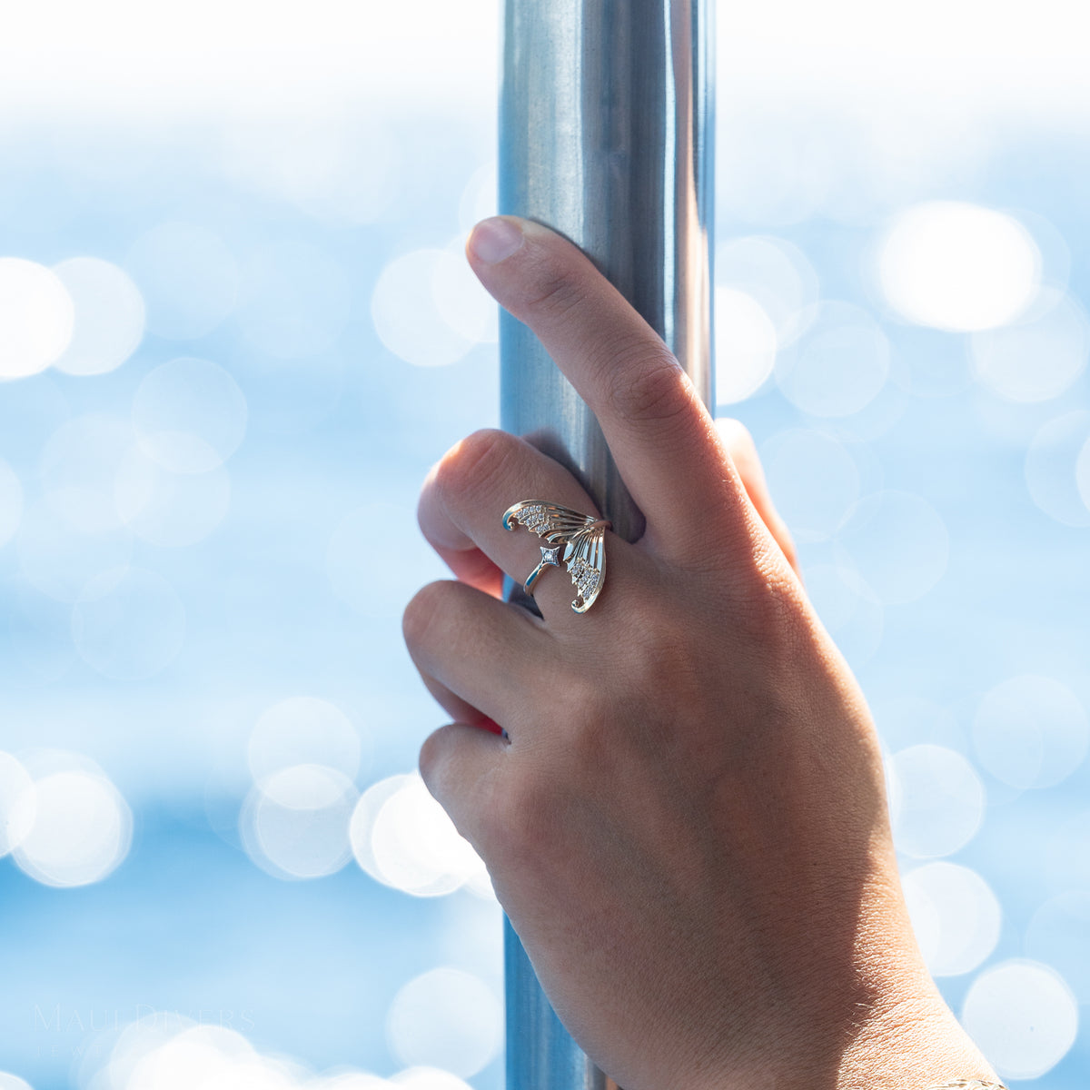 Close-up of a hand wearing a gold bypass mermaid tail ring with diamonds and a white gold diamond star, against a sparkling blue ocean background