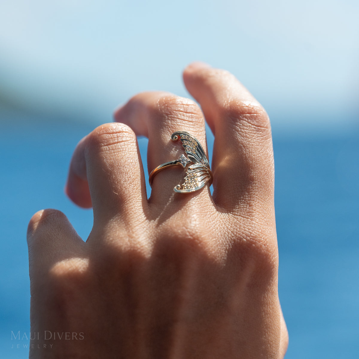 Close-up of a hand wearing a gold bypass mermaid tail ring with diamonds and a white gold diamond star, against a blurry blue ocean background