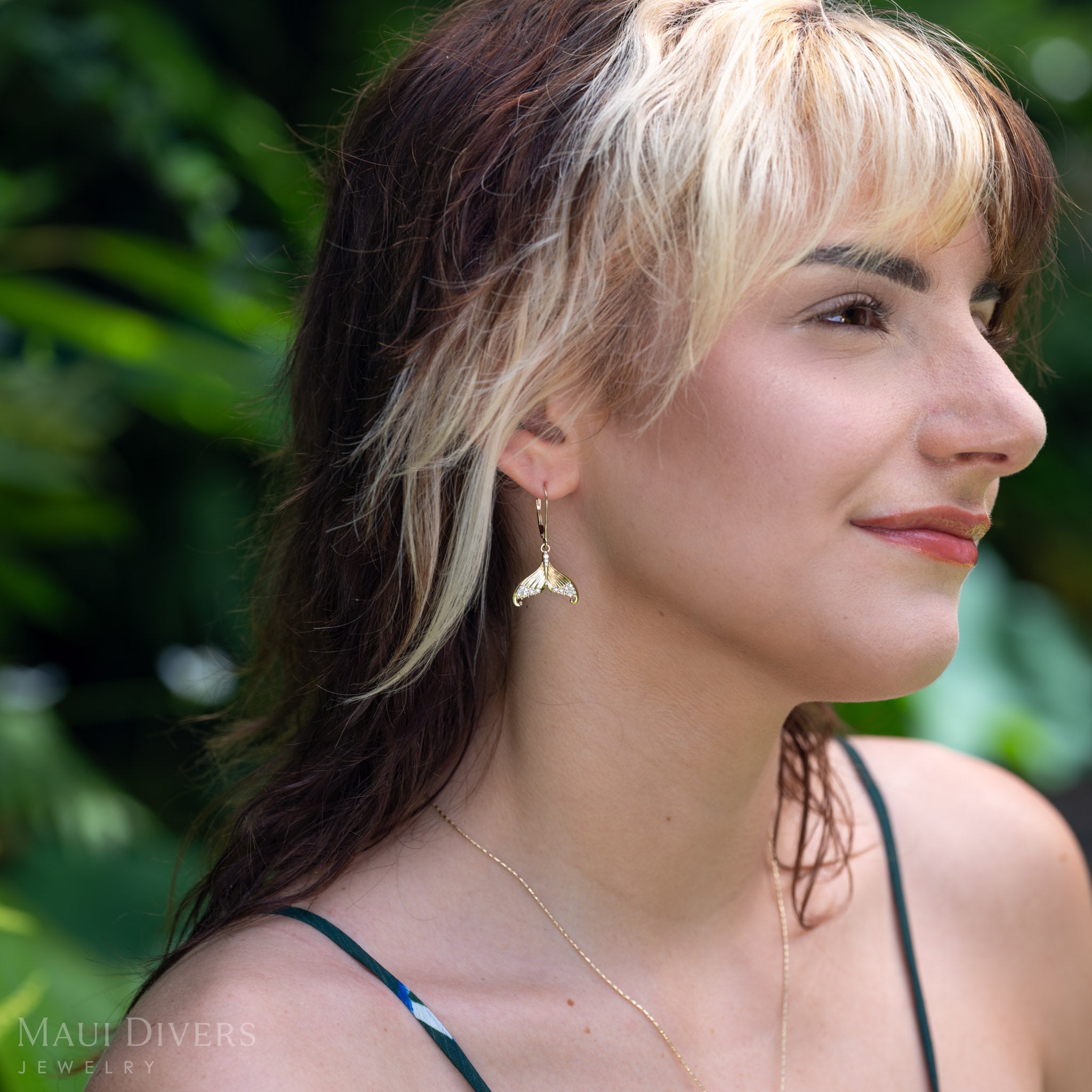 A woman in profile wearing a dangling diamond mermaid tail leverback earring in 14k yellow gold  outdoors with blurred green foliage in the background