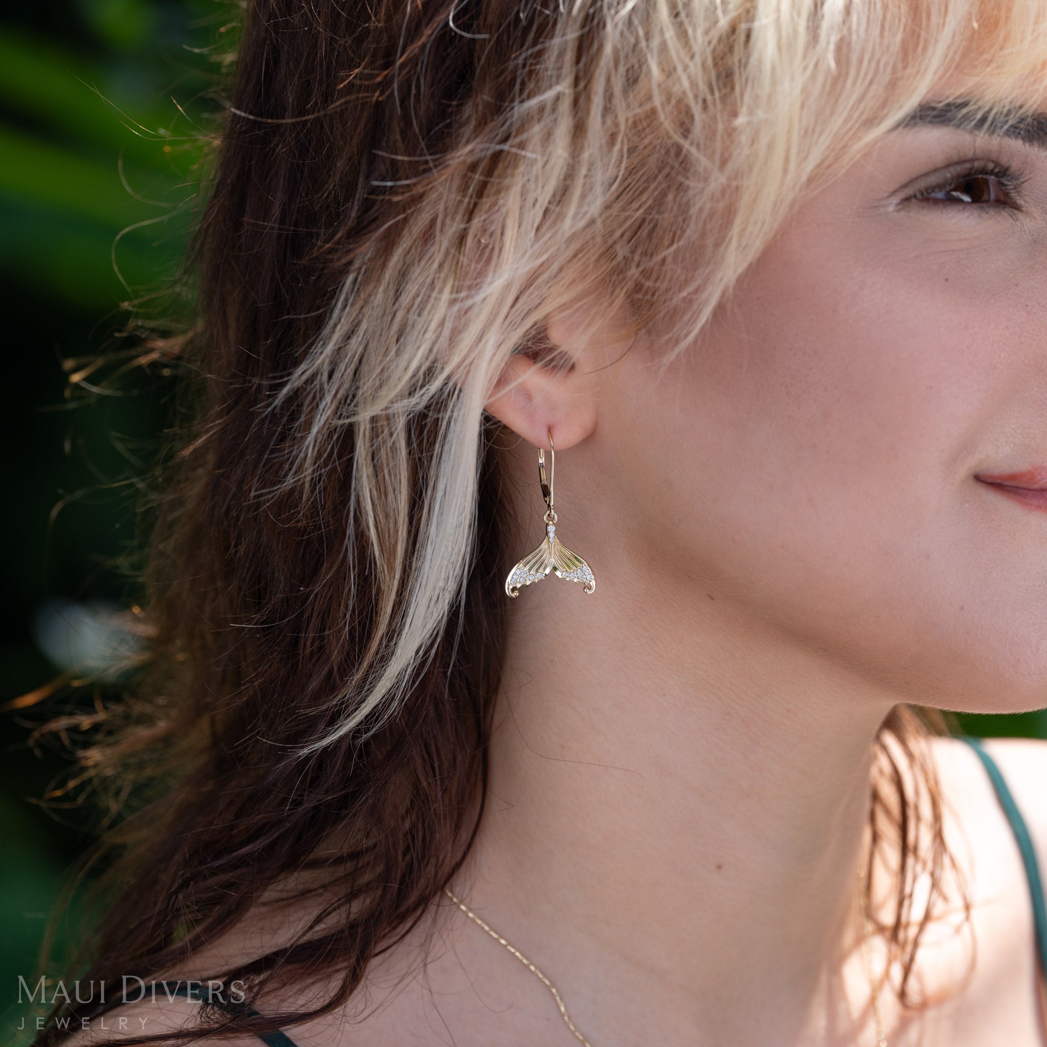 A close-up side profile of a woman with blonde-highlighted hair wearing a dangling diamond mermaid tail leverback earring in 14k yellow gold