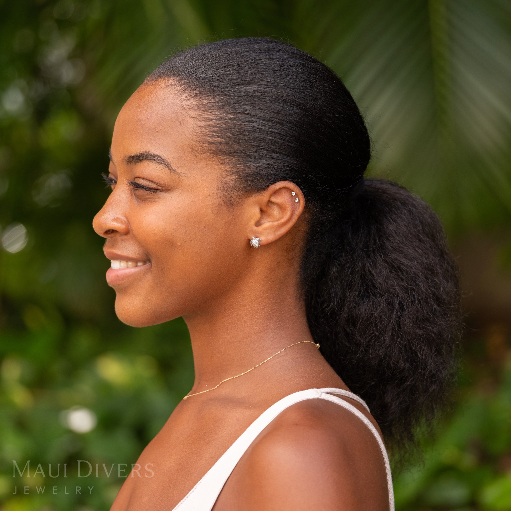 A smiling woman wearing a Honu Earring in 14k White Gold with Diamonds against a blurred leafy green background