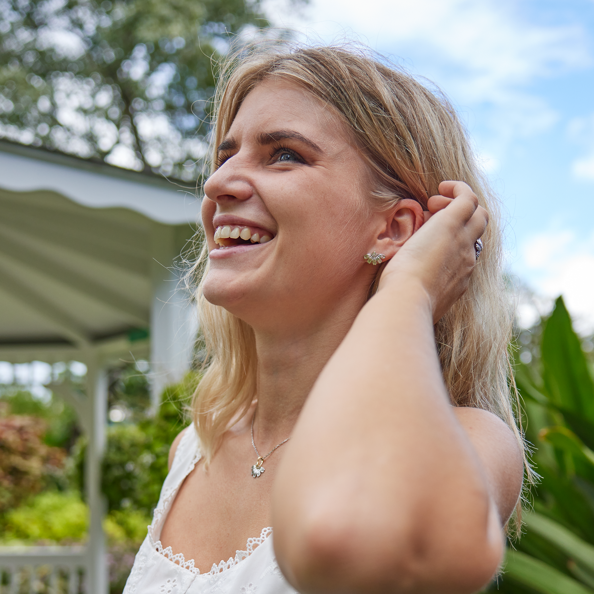 Laughing Woman Wearing Beach Naupaka Earrings in Two Tone Gold with Diamonds with Blue sky and green plants
