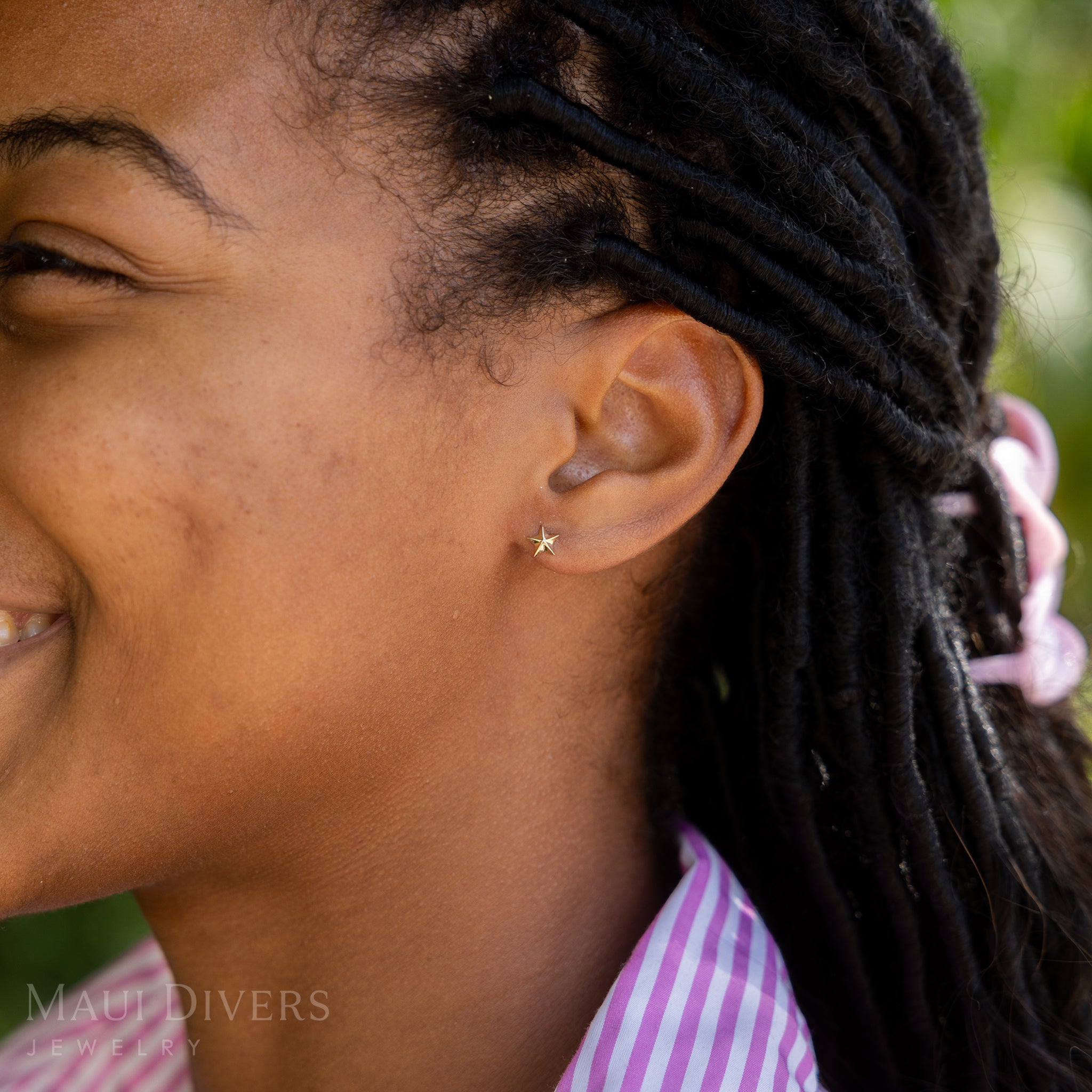 A side view of a smiling woman in a pink and white striped top wearing a Starfish Earring in 14k yellow gold, against a blurred leafy green background