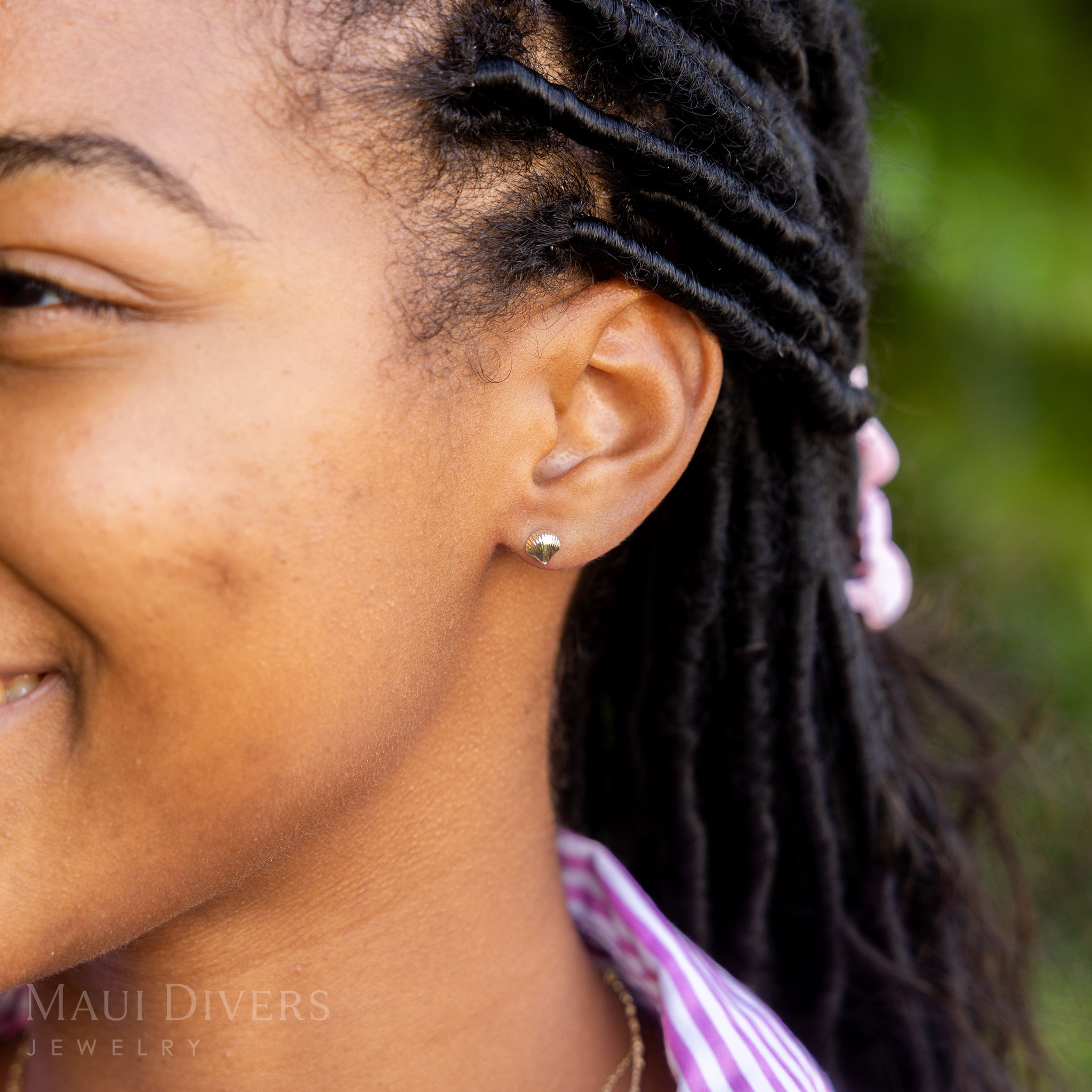 A side view close-up of a smiling woman in a pink and white striped top wearing a Scallop Shell Earring in 14k yellow gold, against a blurred leafy green background