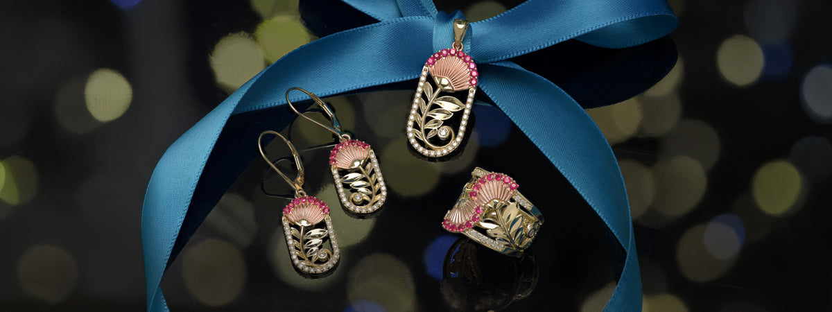 A jewelry set featuring a matching ʻŌhiʻa Lehua flower pendant, earrings, and ring in 14k rose and yellow gold with diamonds and rubies, presented on a glossy black surface wrapped in a blue ribbon against a dark bokeh background.