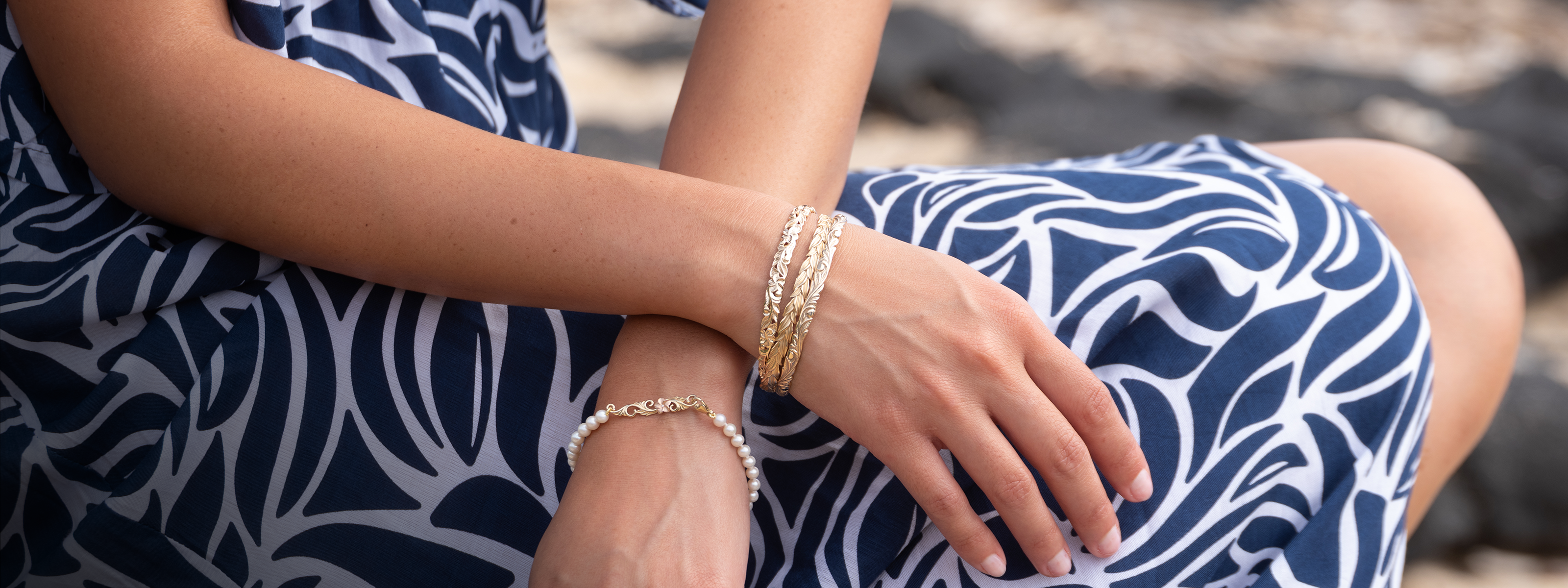 Person wearing a blue and white patterned dress with Hawaiian gold bracelets on a blurred background