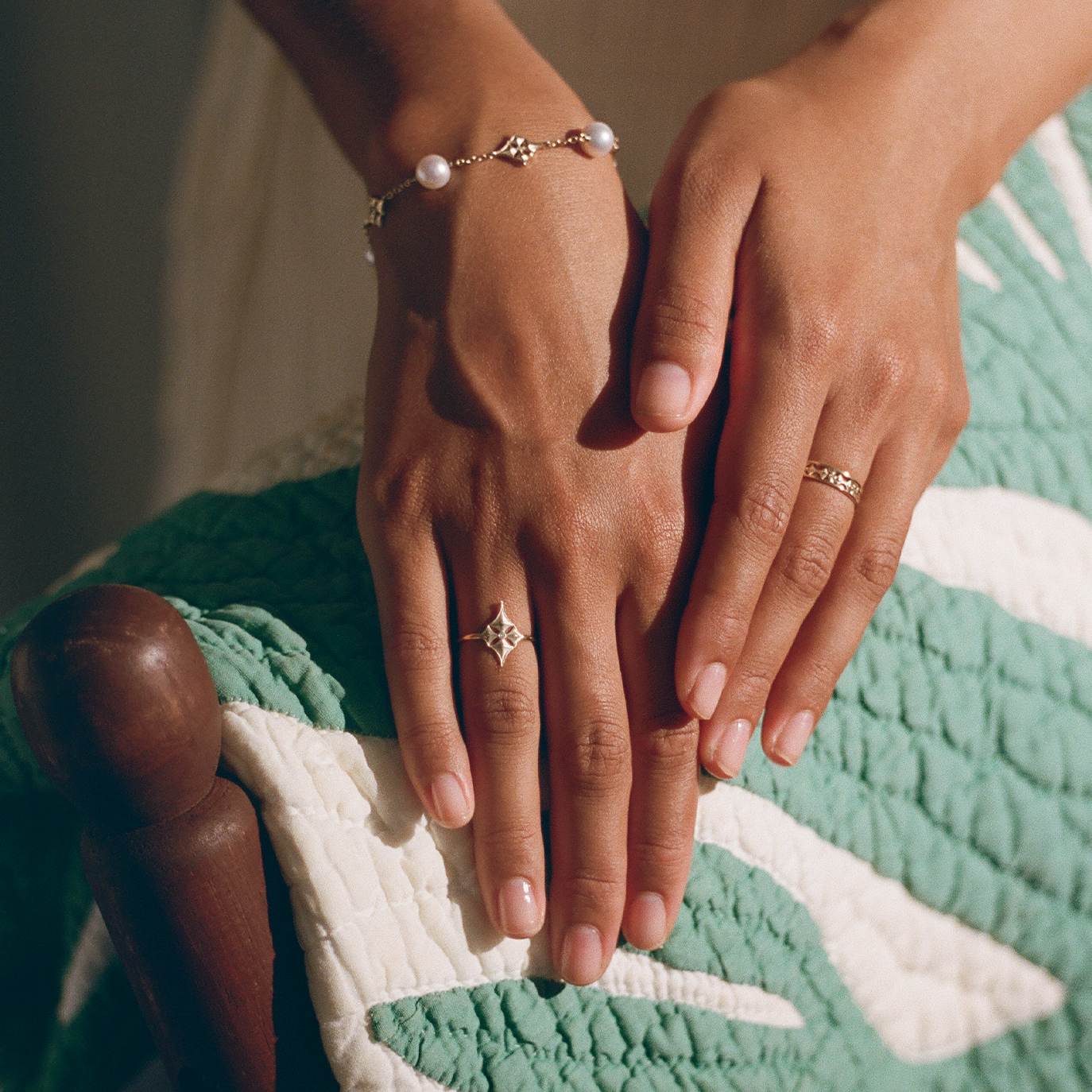 Close-up of hands resting on a green Hawaiian quilt while wearing 14k yellow gold Hawaiian quilted star rings and an Akoya bracelet with alternating Hawaiian quilted stars in 14k gold