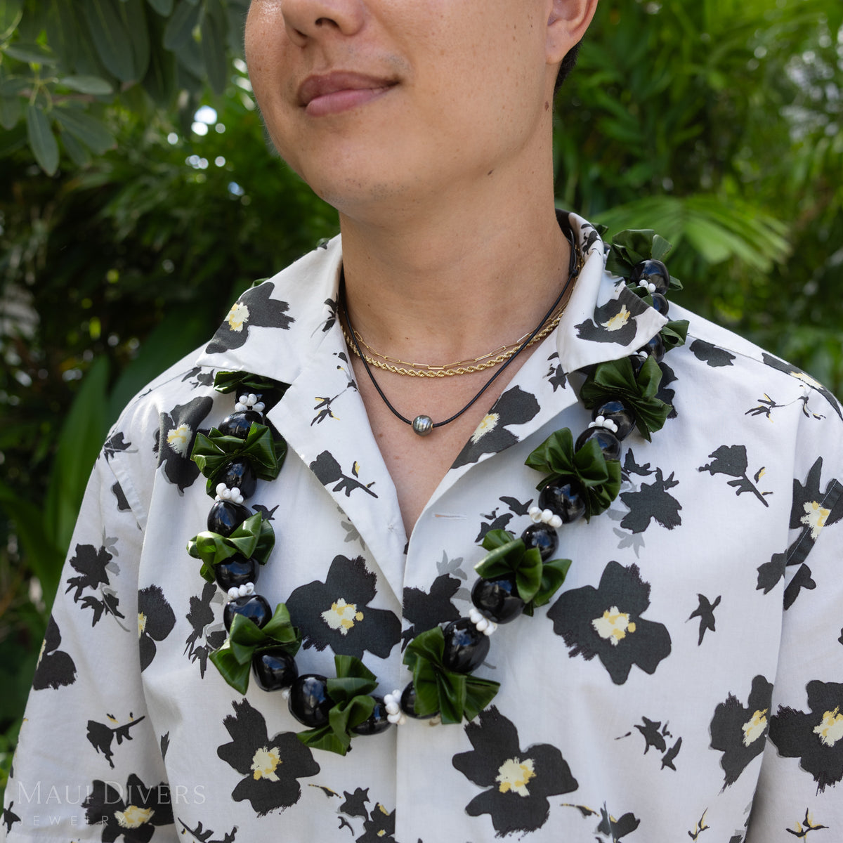 A close-up shot of a man wearing layered gold necklaces and a black coral tahitian black pearl necklace with a lei around him and a patterned white shirt against a blurry nature background