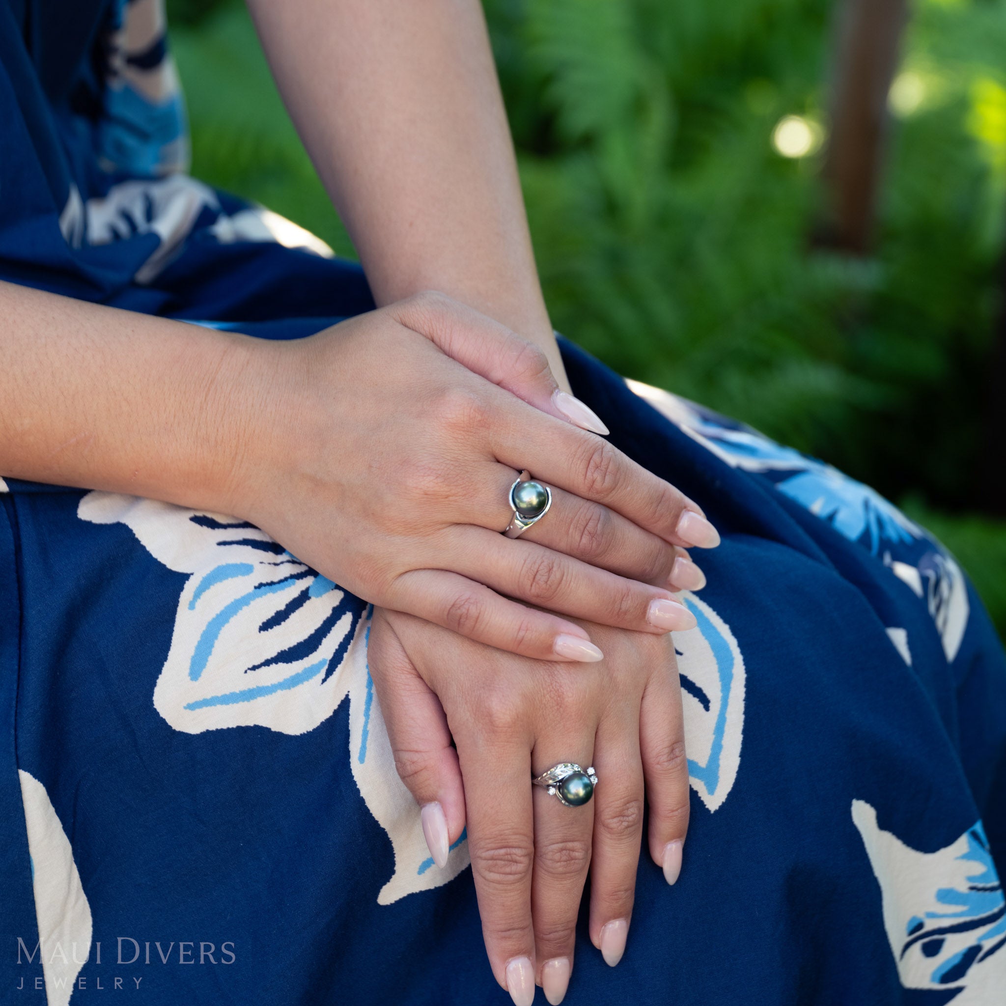 Close-up of hands wearing Tahitian black pearl rings with a blue floral dress and greenery in the background