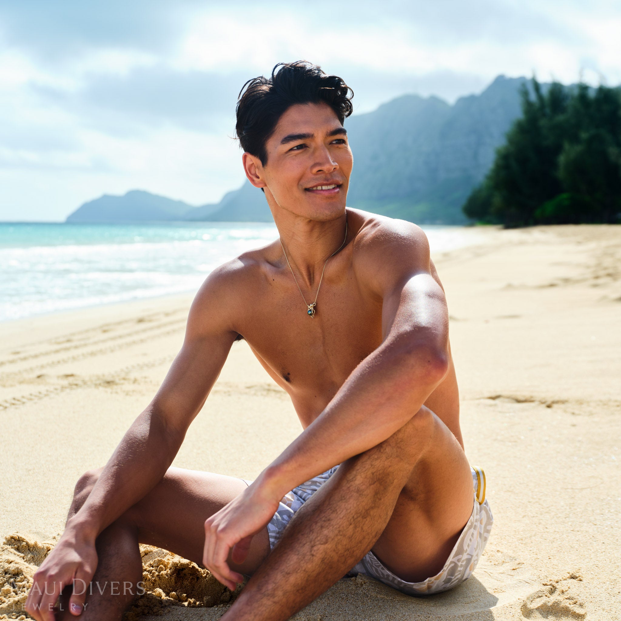 Man sitting on a sandy beach wearing an octopus Tahitian black pearl pendant with mountains and ocean in the background