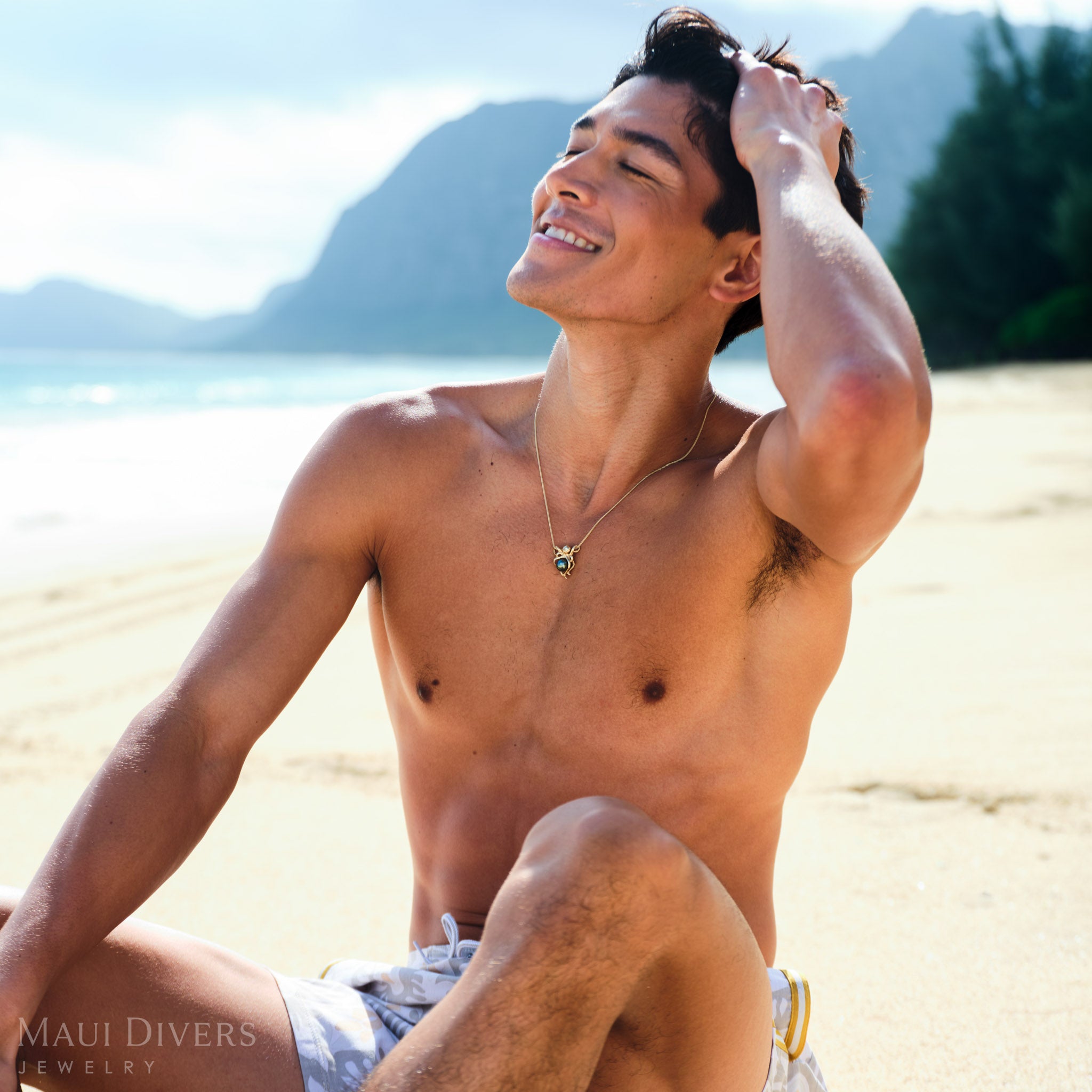 A man sitting on a beach wearing an octopus Tahitian black pearl pendant with mountains and beach in the background
