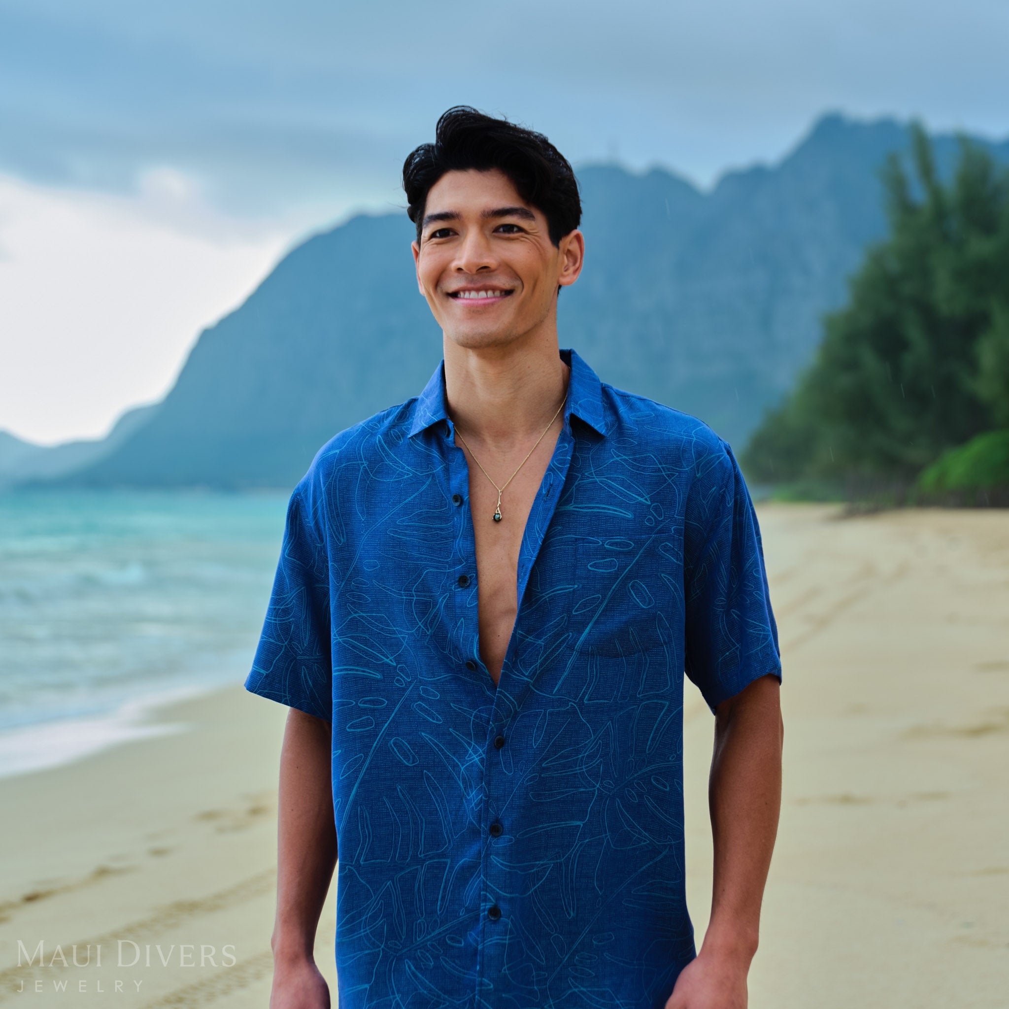 Man wearing gold Tahitian black pearl pendant a blue patterned shirt on a beach with mountains in the background