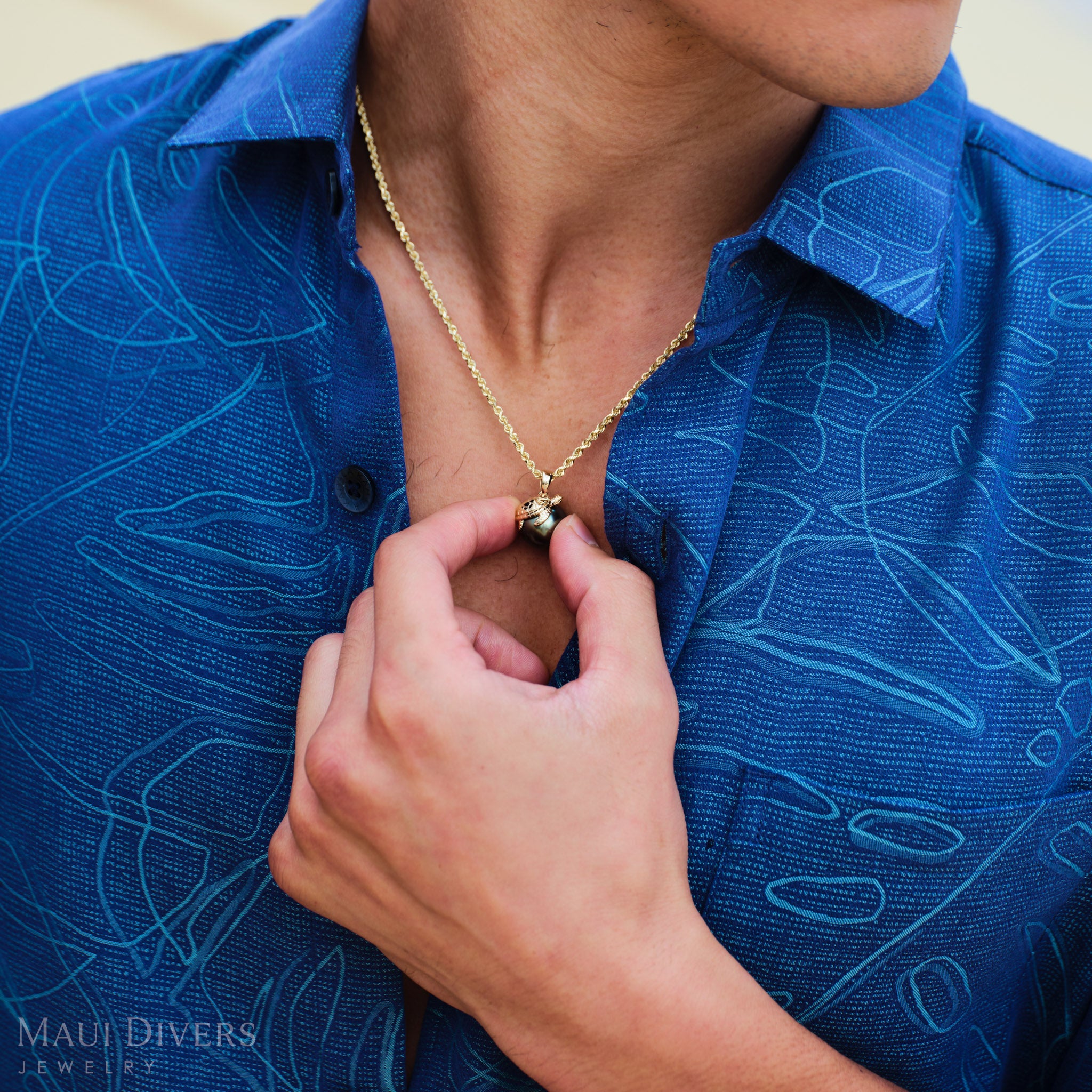 Man wearing a blue patterned shirt and gold necklace, adjusting a sea turtle Tahitian black pearl pendant. 