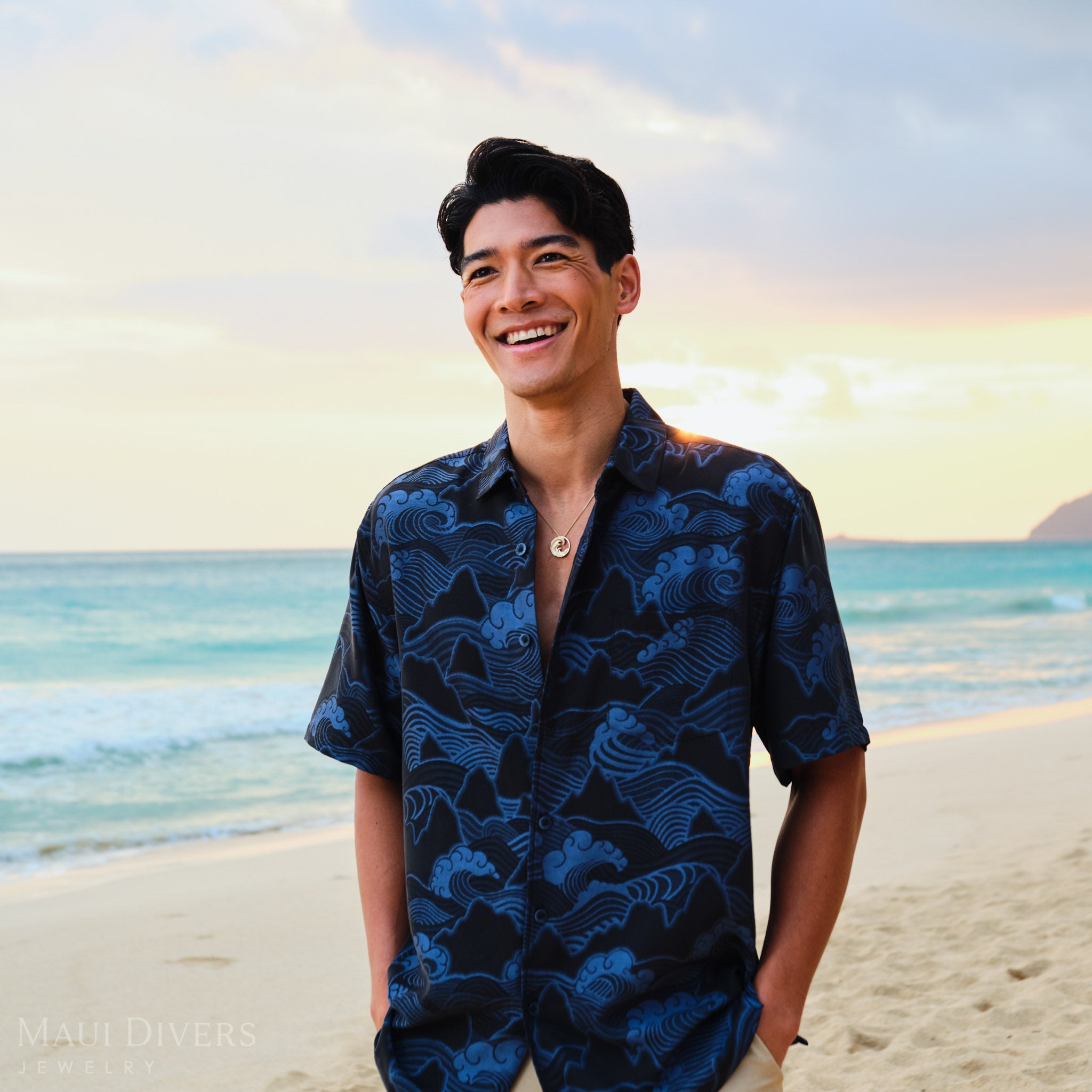 Man on a beach at sunrise wearing a dark blue patterned shirt with a gold diamond wave pendant 