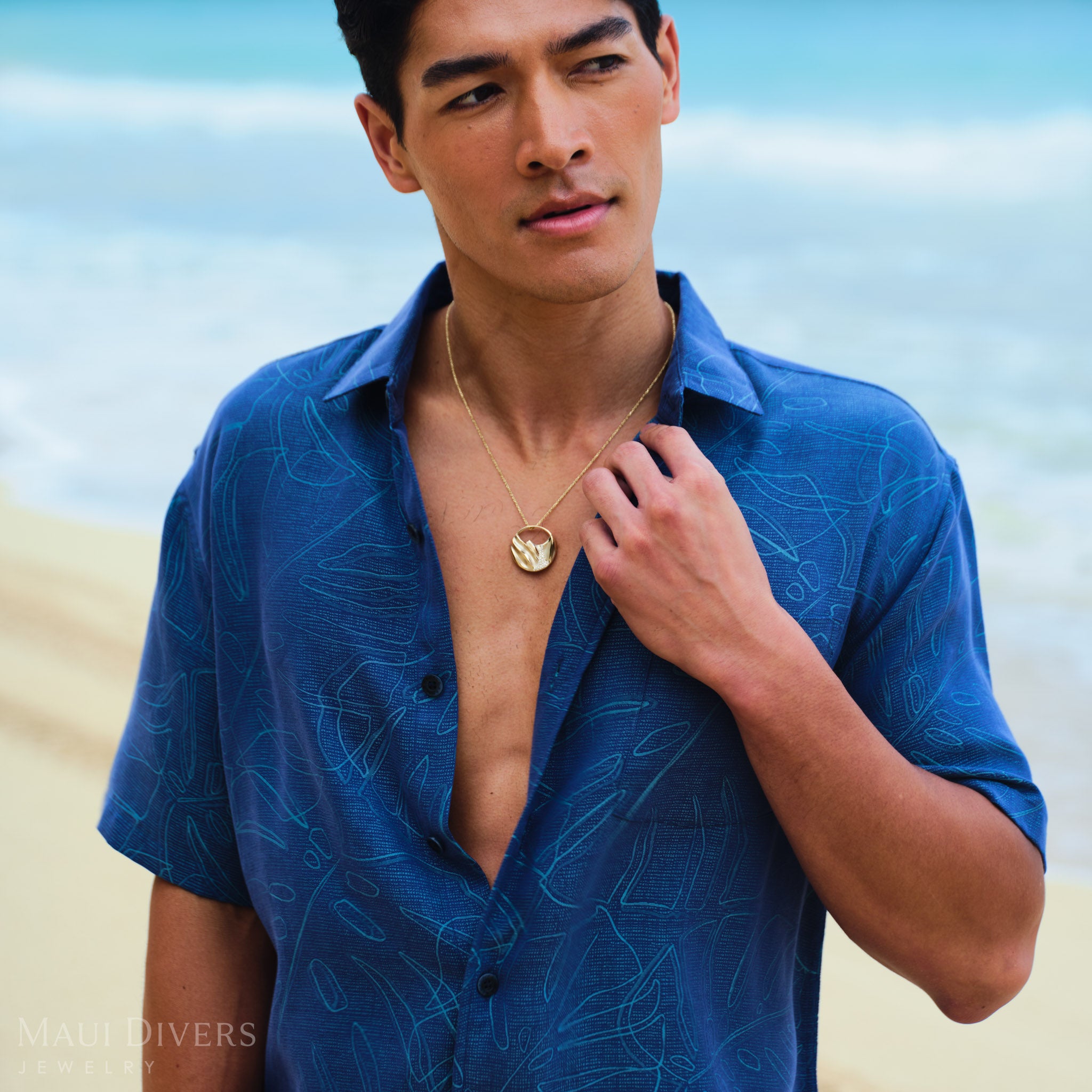 Man wearing a gold Koʻolau mountain pendant and a patterned monstera shirt on a beach