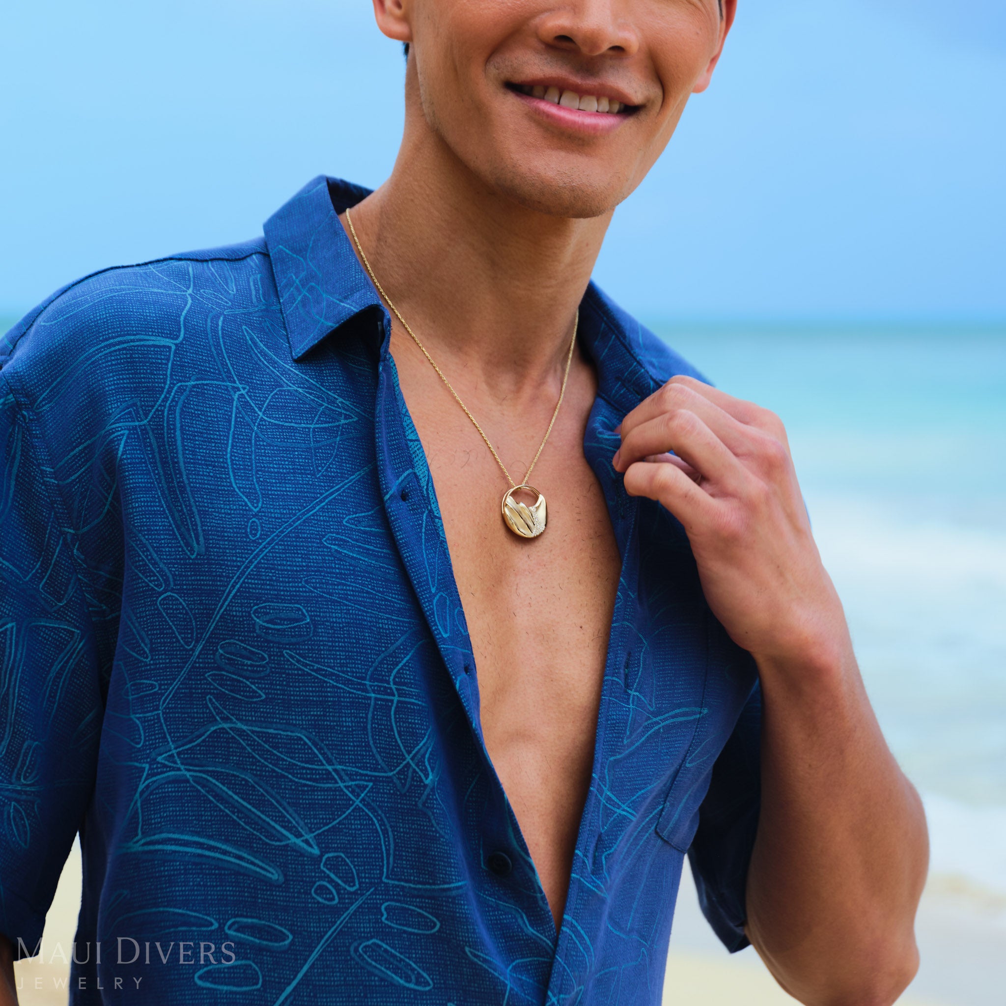 Man wearing a gold Koʻolau mountain pendant and a patterned monstera shirt on a beach