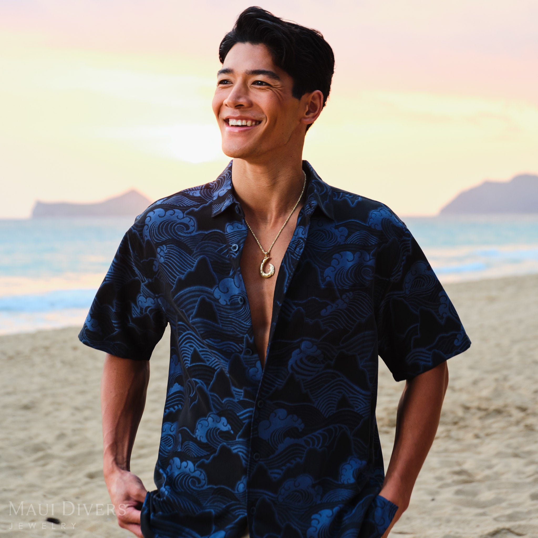 Man wearing a blue patterned shirt and a gold Hawaiian fish hook pendant on a beach with a sunrise in the background